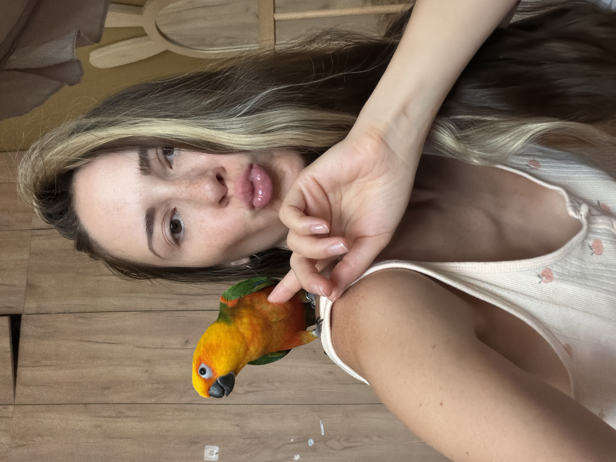 A medium shot shows a young woman with long, brown hair with blonde highlights, and freckles on her nose and cheeks. She is wearing a white tank top with small pink fruit patterns. The woman is making a kissy face with her lips puckered and her right hand is near her chin, with her index finger and thumb forming a "pinch" gesture. A small, vibrantly colored parrot, predominantly orange and yellow with green accents, is perched on her left shoulder. The background appears to be a wooden wall or closet doors. There is also a blurred object with a rabbit ear shape visible to the right of the woman. The image does not provide any specific information or visual cues to indicate its location in Khimki, Russia.
