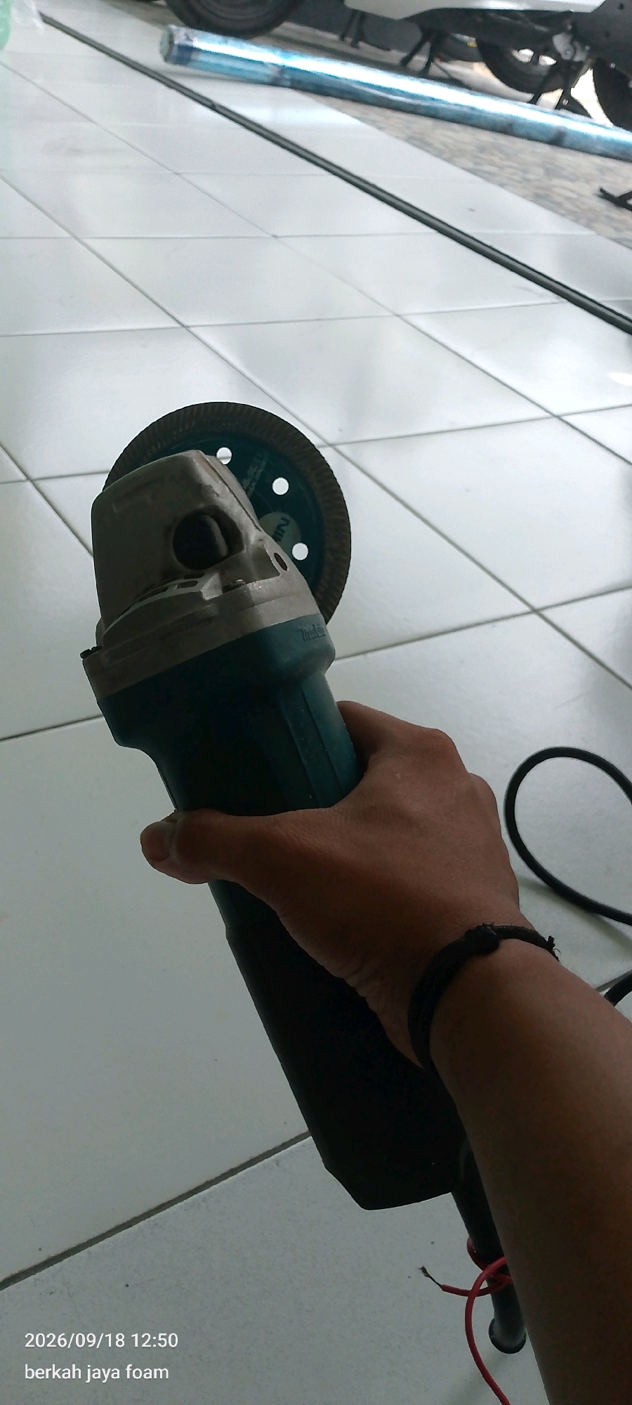 A person's hand is holding a teal and silver angle grinder with a circular cutting disc attached. The grinder is positioned diagonally, with the cutting disc facing the viewer. The hand is wearing a black wristband and is gripping the body of the angle grinder. The background consists of a tiled floor with white square tiles and a black line running across them. In the upper portion of the image, a motorcycle and a blue rolled-up object are visible. The bottom left corner of the image displays a timestamp "2026/09/18 12:50" and text "berkah jaya foam". The overall setting appears to be indoors, possibly a workshop or garage, with a white tiled floor. The location is specified as Kincang Wetan, Indonesia.