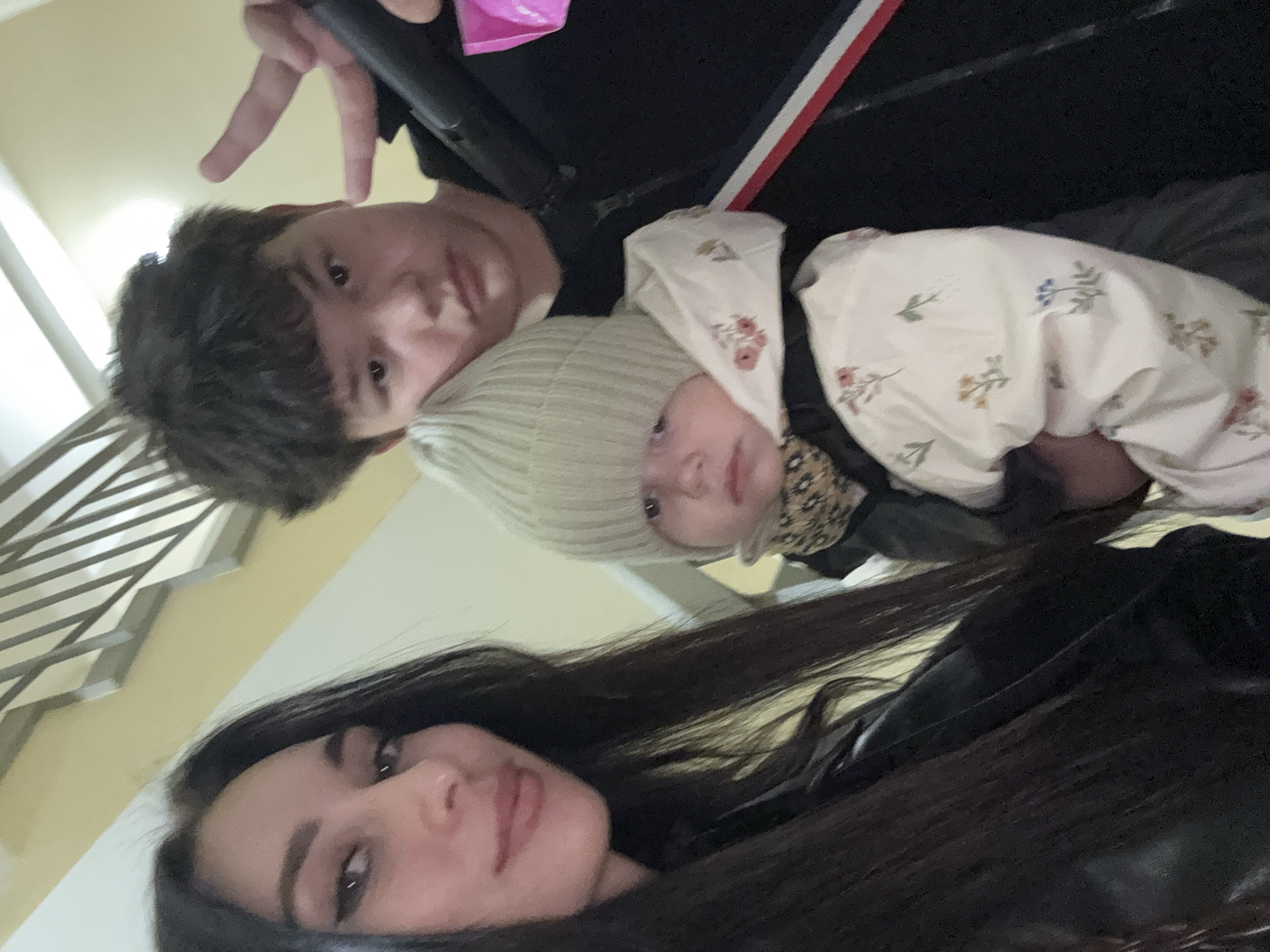 A selfie taken indoors features three individuals: a woman with dark hair and visible makeup (eyeliner and lip gloss), a young man with dark, short hair, and an infant wearing a light-colored floral-patterned hooded outfit and a striped beanie. The background appears to be an interior staircase area with a light-colored wall. A hand making the "peace sign" is visible above the young man. The precise location in the city of Chaikovsky, Russia, is not discernible from this image alone.