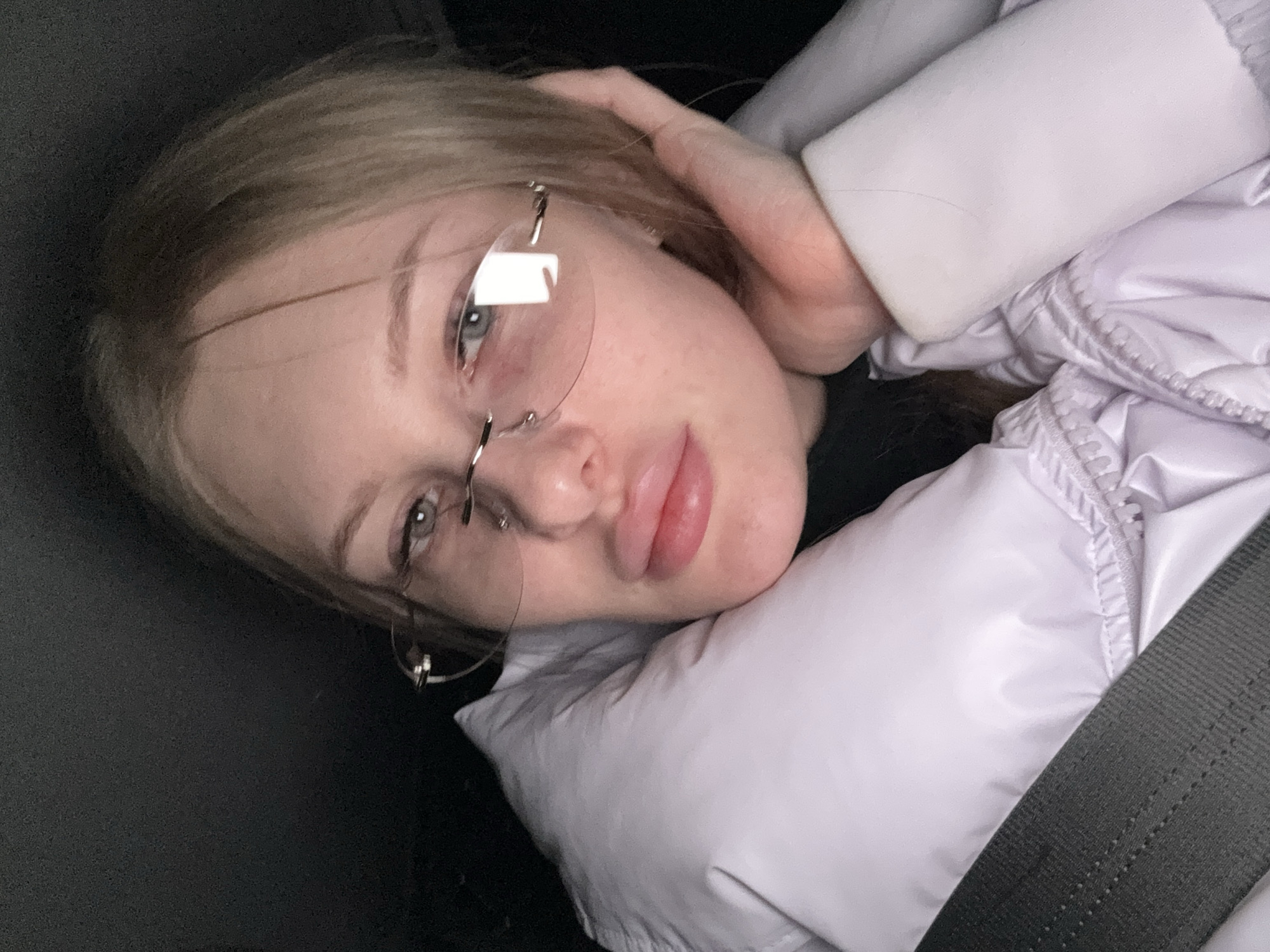 A young woman with fair skin and blonde hair is positioned in a vehicle. She is wearing rimless glasses. The woman is wearing a light purple puffy jacket. The woman's head is resting on a puffy material. A seatbelt is visible in the frame. The location is unknown and there is no information to suggest it is Perm, Russia.