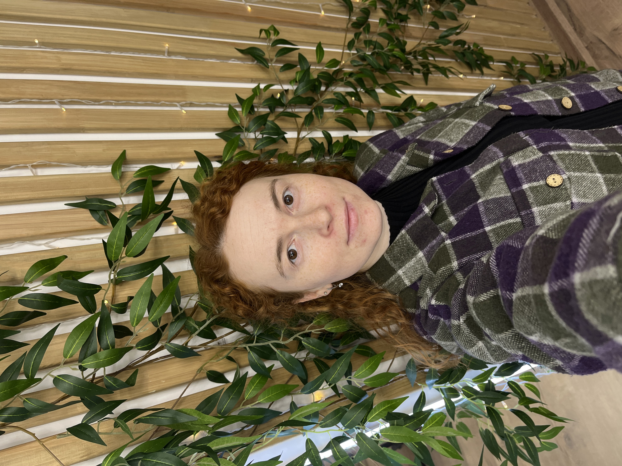 This rotated close-up portrait features a person with reddish-brown, curly hair, fair skin, and freckles, looking directly at the camera with a neutral expression. Their head is positioned towards the lower-left of the frame. They are wearing a plaid jacket or overshirt in shades of dark green, purple, and white, adorned with small wooden buttons, over a black ribbed high-neck top. Small, silver-toned earrings are visible in their left ear.
The background is composed of light-colored, horizontal wooden slats. Artificial green foliage with slender leaves is decoratively draped across the wooden panels, intermingled with delicate string lights that emit a soft, warm glow. The arrangement creates a natural and cozy aesthetic, suggesting an indoor setting or a specially decorated area. The image appears to be taken in Chaikovsky, Russia. No text is visible.