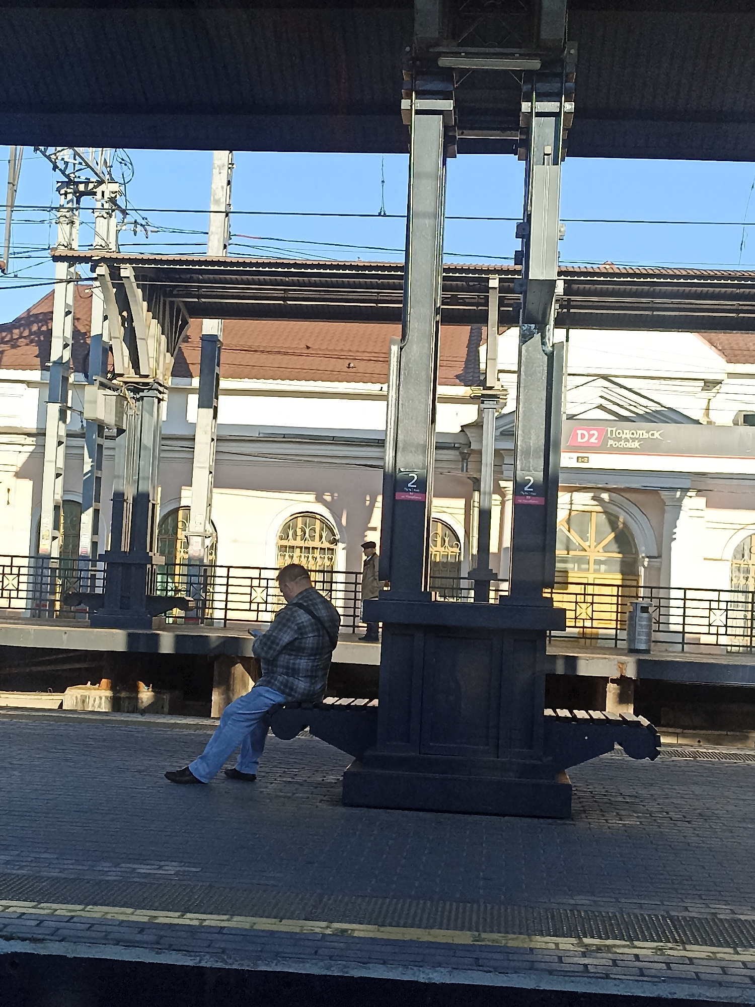 This is a full-frame, eye-level, outdoor photograph taken on a sunny day at a train station in Podolsk, Russia. In the foreground, a man wearing a plaid shirt and blue jeans is sitting on a bench and looking down at his phone. The bench is part of a dark-colored, modernistic structure with thick, vertical supports. Behind the man, a train platform extends to the left and right. A building with light-colored walls and a reddish-brown roof is visible in the background, featuring arched windows. Overhead, there is a canopy supported by metal beams, and complex electrical infrastructure for the trains is apparent. A sign on the building reads "D2 Подольск Podolsk". The ground is paved with dark tiles and yellow safety lines. The overall scene depicts a typical urban train station environment.