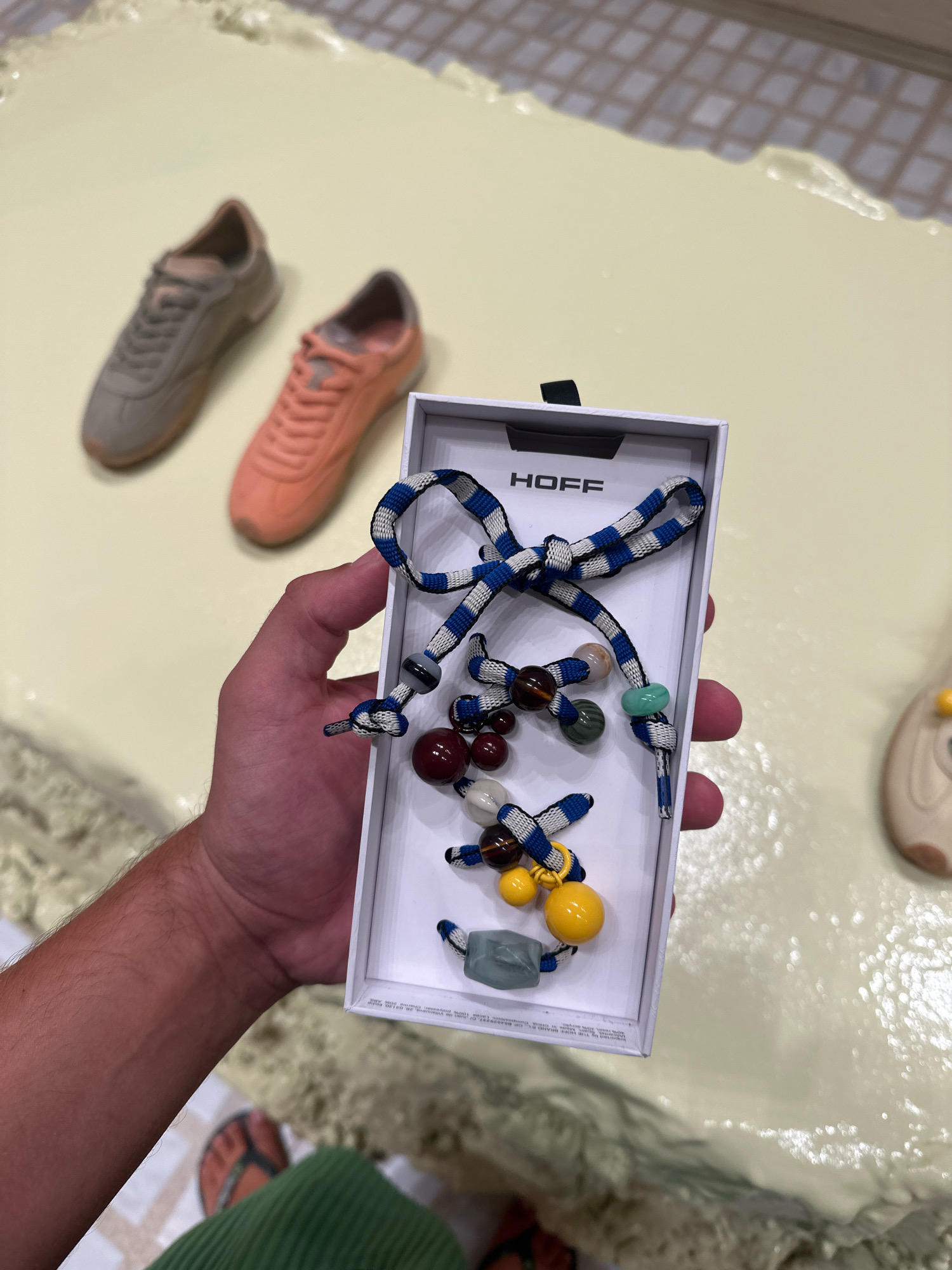 The image shows a person's hand holding a white box containing a blue and white striped cord with various beads attached, resembling a bracelet or keychain. The box has the logo "HOFF" printed on it. In the background, two blurred sneakers, one grey and one peach, are visible on a light-colored surface. The environment appears to be indoors, possibly a store or a personal space. There is no indication that this scene is located in Madrid, Spain.