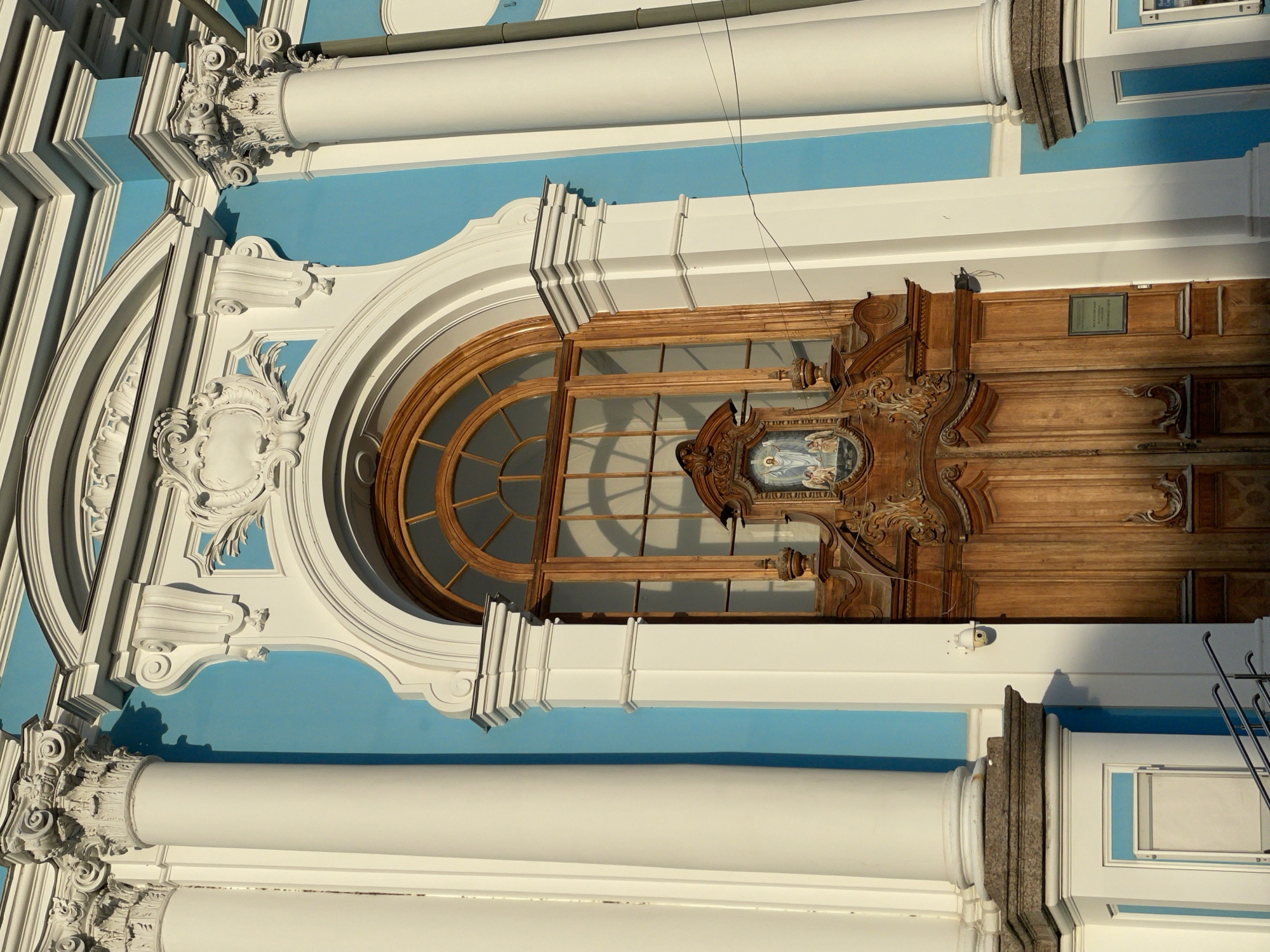 This is a detailed, slightly upward-angled view of the highly ornate main entrance facade of the Smolny Cathedral in St. Petersburg, Russia. The building features a vibrant light blue background color for its walls, contrasted by stark white architectural details.
Two massive white Corinthian columns, with fluted shafts and intricately carved capitals, frame a prominent central arched entryway. The columns exhibit distinct shadows cast onto the blue wall behind them, indicating strong directional sunlight from the left.
The archway itself is framed by layers of elaborate white moldings, showcasing Baroque or Rococo stylistic elements. Within this arch, a tall, double wooden door is present, made of light brown wood. The door's upper panels are richly carved with volutes, scrolls, and decorative patterns. Centered above the functional door panels, an embedded religious icon or painting depicts a standing figure, presumably Christ, flanked by two angels in an ethereal setting, all within an ornate wooden frame.
Above the wooden door, and integrated into the same arched opening, is a large arched window with a wooden frame and a geometric grid of mullions. Reflections of unseen structures are visible on the windowpanes.
Above the main archway, against the blue wall, are more elaborate white decorative elements, including a central cartouche surrounded by scrollwork and foliate designs.
To the right of the wooden door, on a white pilaster, a small white security camera is affixed. A dark rectangular plaque with white text is mounted on the lower right section of the door's frame. In the lower left corner, partial metal handrails of a staircase are visible, suggesting access steps to the entrance. A dark vertical utility pipe runs along the rightmost white column.
No individuals are visible in the scene. The overall impression is one of grandeur and intricate architectural detail under clear, bright lighting.