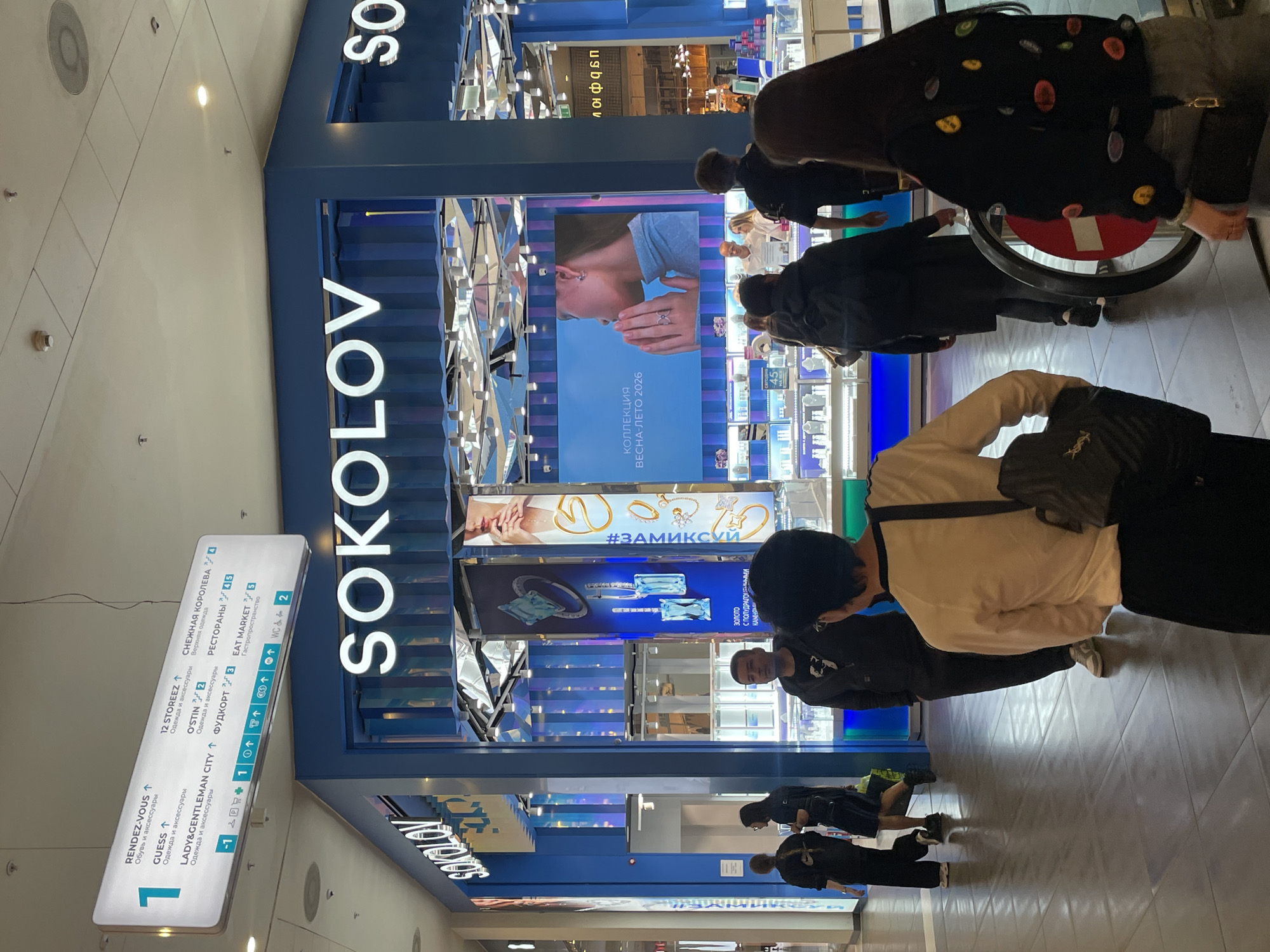 The image shows the exterior of a "SOKOLOV" jewelry store, likely in a shopping mall, in Saint Petersburg, Russia. The store's name is prominently displayed in large, illuminated letters. Several people are seen browsing the window displays, and one person is in a wheelchair. A directory sign lists various stores and restaurants, including "RENDEZ-VOUS," "GUESS," "12 STOREEZ," "O'STIN," "LADY&GENTLEMAN CITY," "СНЕЖНАЯ КОРОЛЕВА" (Snow Queen), "РЕСТОРАНЫ" (Restaurants), and "EAT MARKET". The store's windows display various jewelry items with promotional banners, including "#ЗАМИКСКИ" and "КОЛЛЕКЦИЯ ВЕСНА-ЛЕТО 2026" (Collection Spring-Summer 2026). The environment is well-lit and appears to be a busy shopping area.
