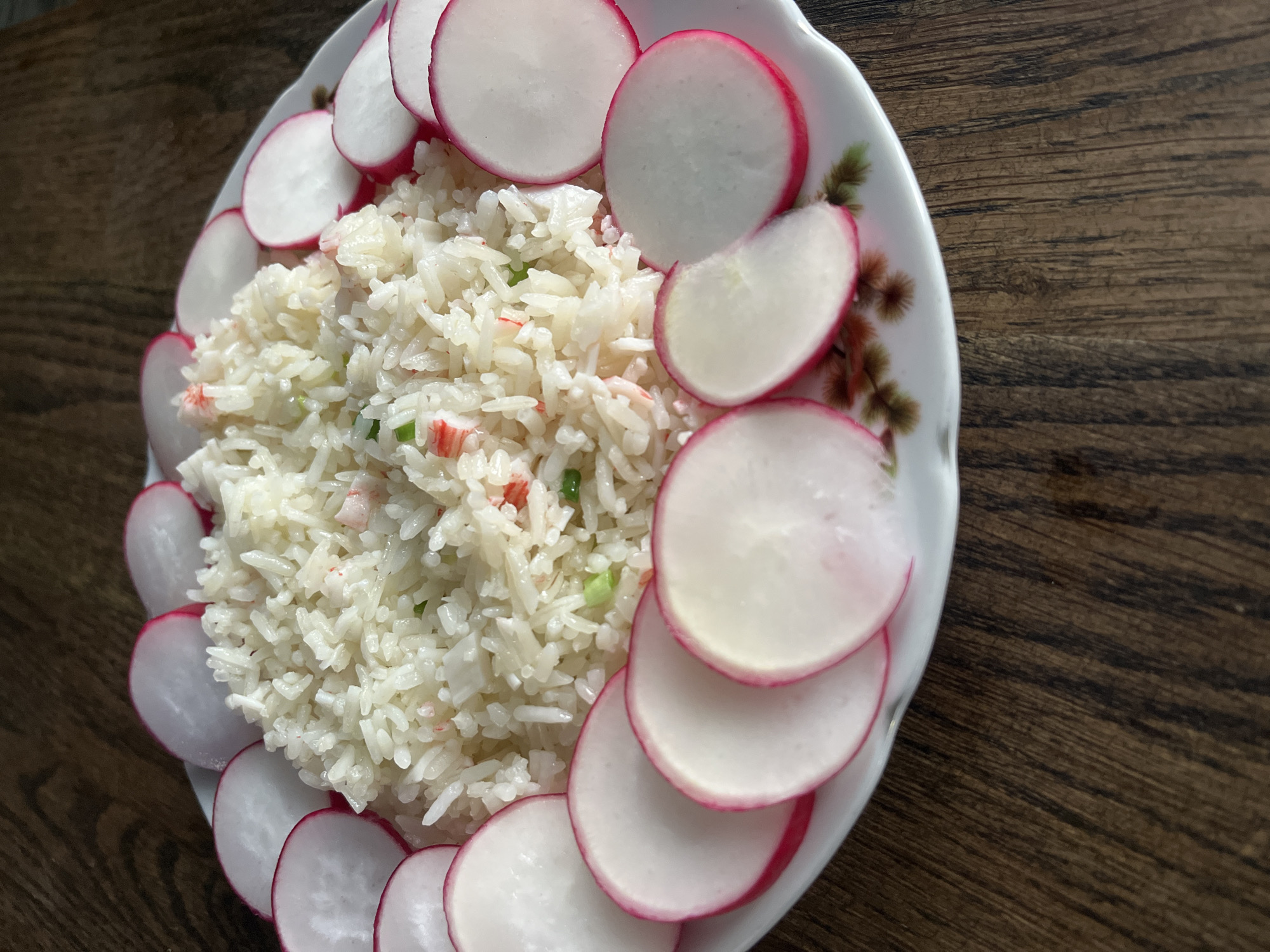 The image shows a plate of rice garnished with sliced radishes, resting on a dark wood-grain surface. The rice appears to contain small pieces of green and red ingredients. The radishes are sliced into thin, circular pieces, arranged around the edge of the rice. The plate has a decorative border with a floral pattern. The setting appears to be indoors. The image provides no information about Krasnodar, Russia.