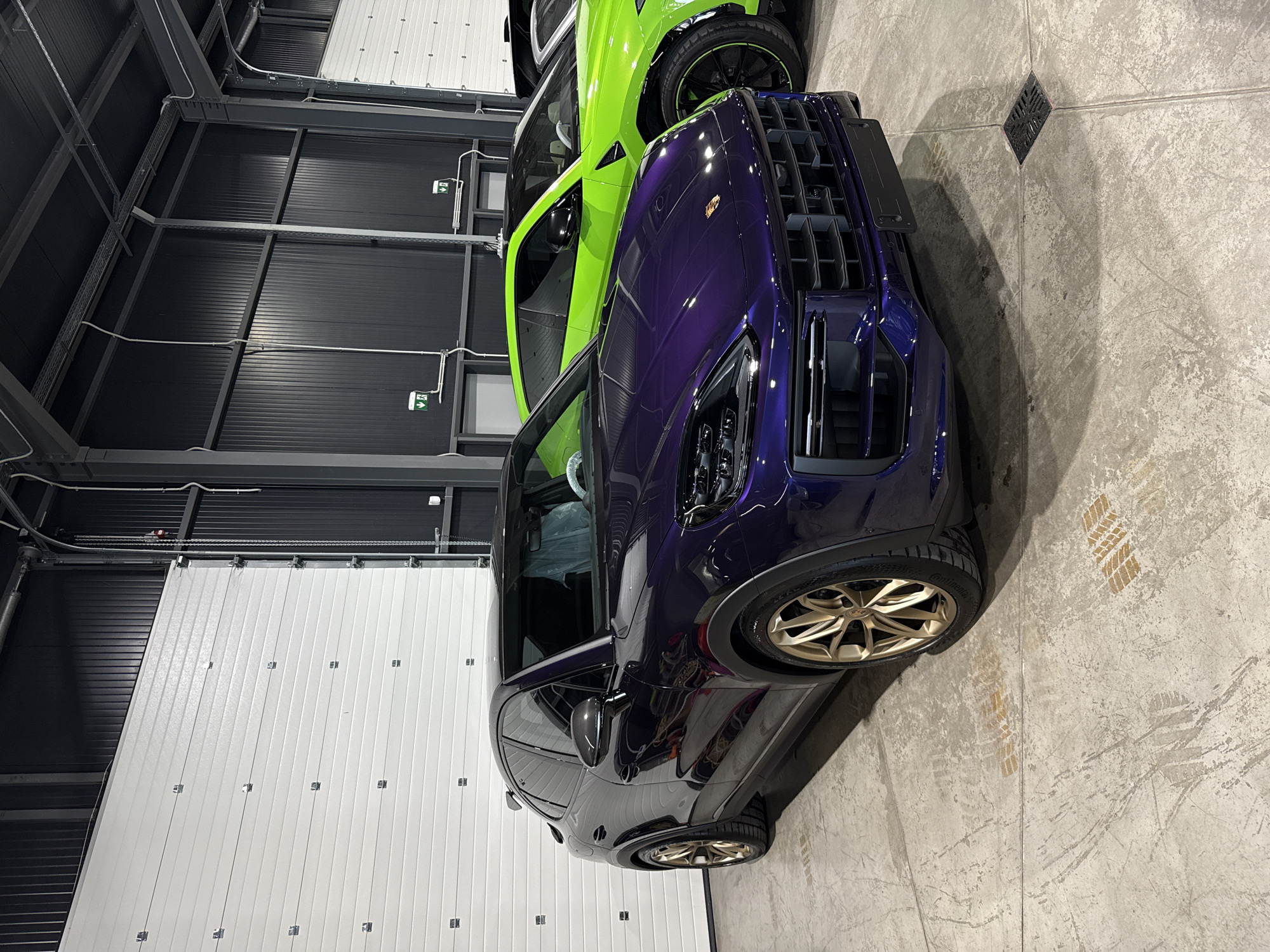 The image shows two cars parked in what appears to be a garage or storage area. The car in the foreground is a dark purple Porsche SUV with gold-colored rims. Next to it is a lime green SUV. The floor is concrete. The walls consist of white sectional garage doors on the left and black corrugated metal walls above. The location is likely Plovdiv, Bulgaria, because no other location was specified.