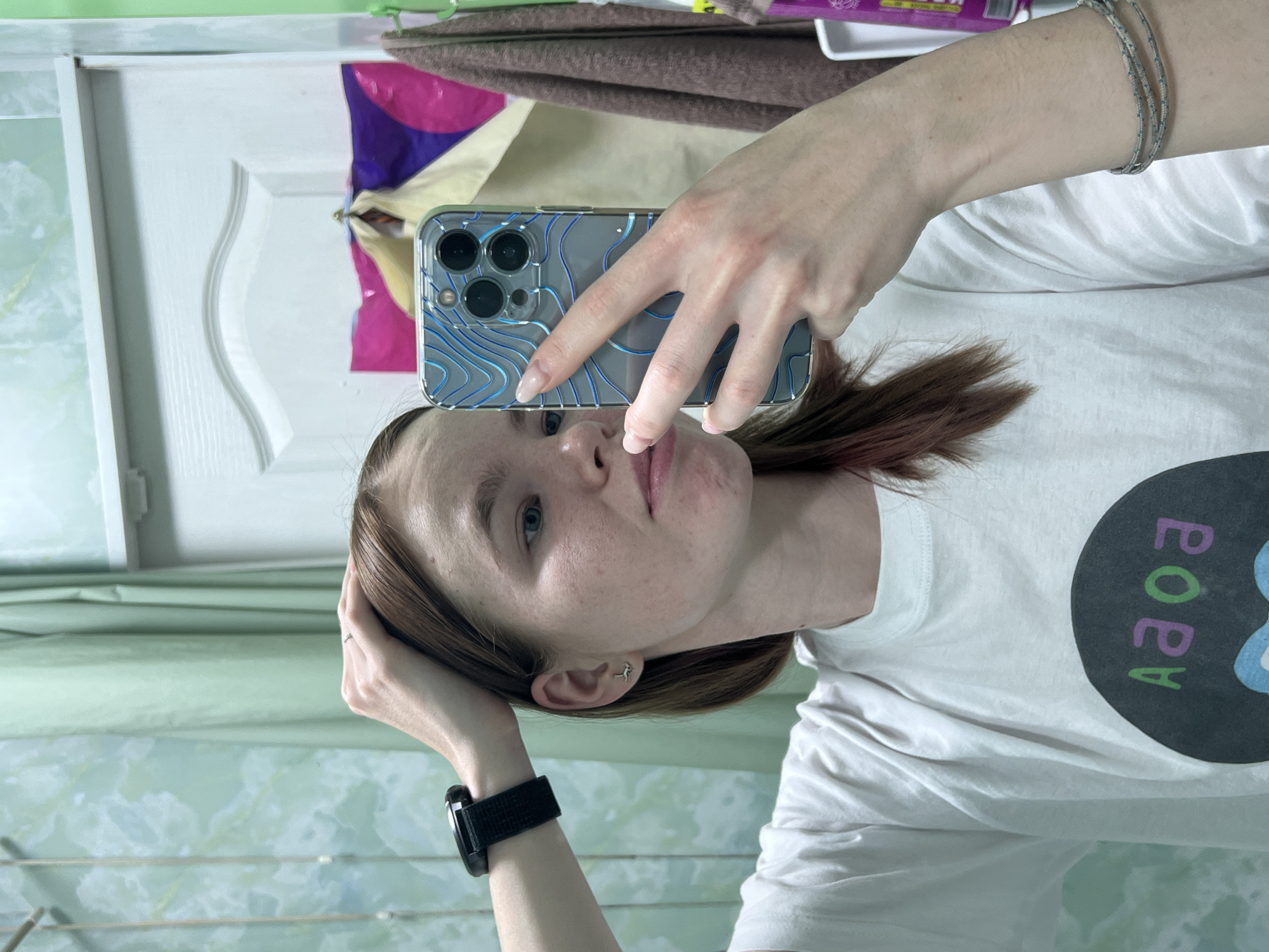 A person with fair skin and reddish-brown hair is taking a selfie in what appears to be a domestic interior. They are wearing a white t-shirt with a circular graphic containing letters in a non-Latin alphabet, and a black watch on their left wrist. The phone case covering the smartphone has a blue topographic pattern. The background includes a white door and a light green wall, possibly in the city of Chaikovsky, Russia. Other details in the background include a pink and purple object, a brown towel and a white object. The selfie style suggests the person is the primary subject of the image.