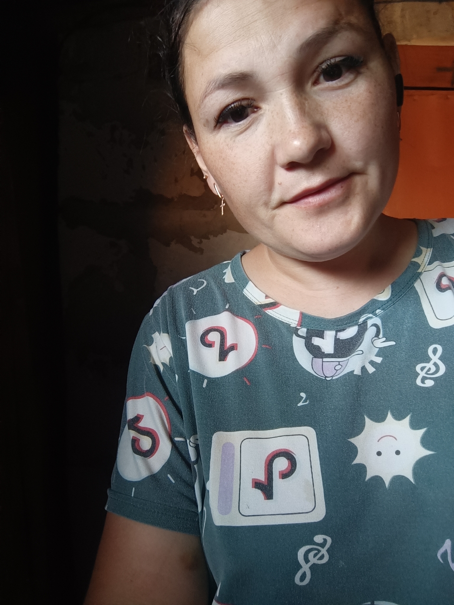A selfie of a woman of East Asian descent is taken from a slightly low angle. She has dark hair pulled back and is wearing a teal t-shirt with a pattern of cartoonish symbols and numbers. She has light brown eyes, full lips, and is looking towards the camera with a slight smile. She has freckles across her nose and cheeks. She is wearing a small gold cross earring on her left ear. The background is dimly lit and appears to be an interior space with dark walls and an orange-colored surface on the right. There is no visible indication of the city of Karsha, Russia.
