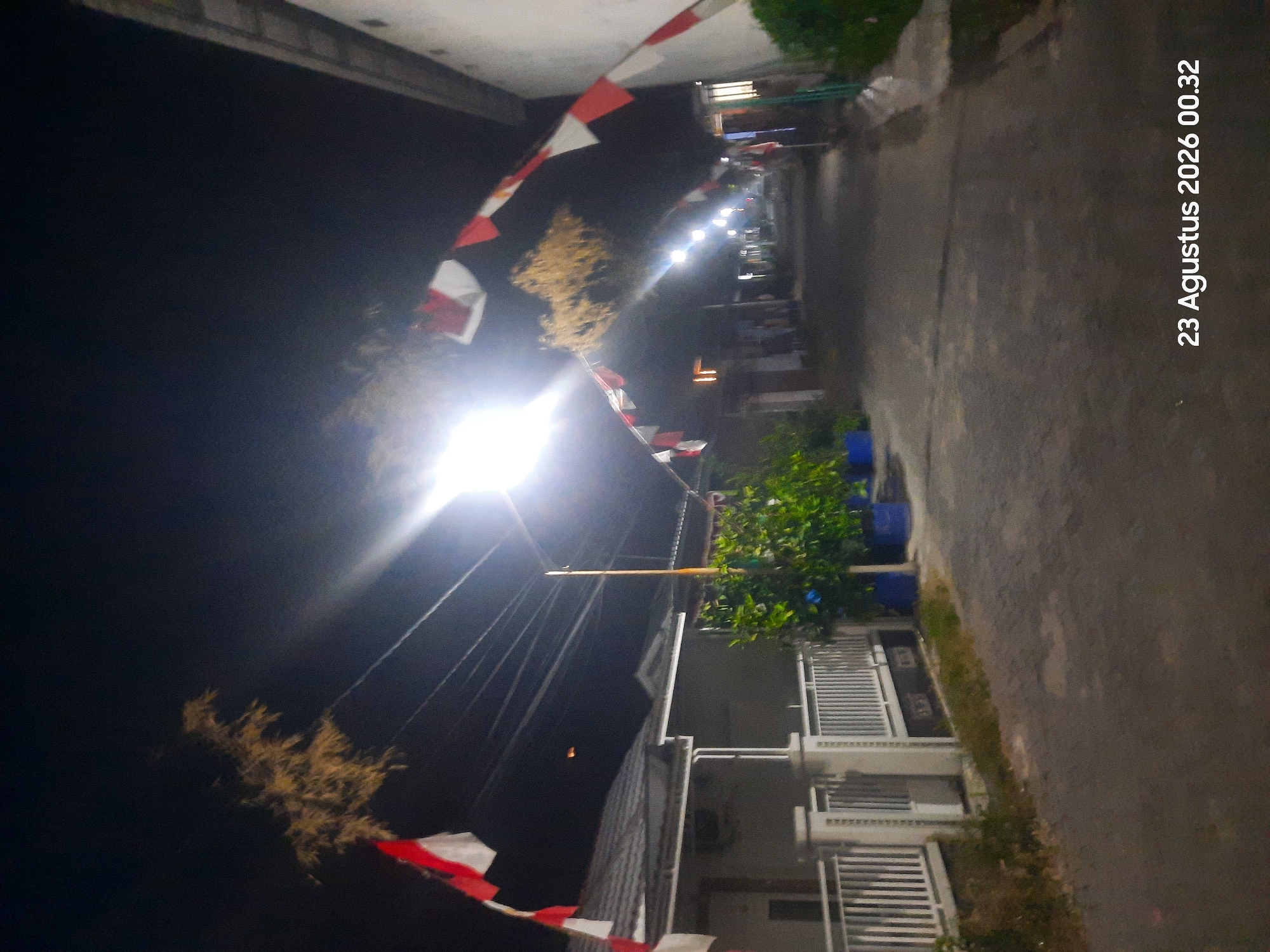 An exterior, night-time, street-level shot shows a residential street in Sukokerto, Indonesia, decorated with a string of red and white Indonesian flags. The flags are strung across the street and attached to poles and buildings, indicating a possible celebration or national holiday. A brightly lit streetlamp illuminates the scene, casting long shadows. The street is paved with interlocking concrete blocks. On the left side of the street, a white fence and the facade of a house are visible, with some greenery in pots. On the right side, a row of houses and more greenery can be seen. The overall atmosphere is quiet and dimly lit, typical of a residential area at night. The date and time "23 Agustus 2026 00.32" are overlaid in the bottom right corner of the image, suggesting this is a timestamp of the photograph.
