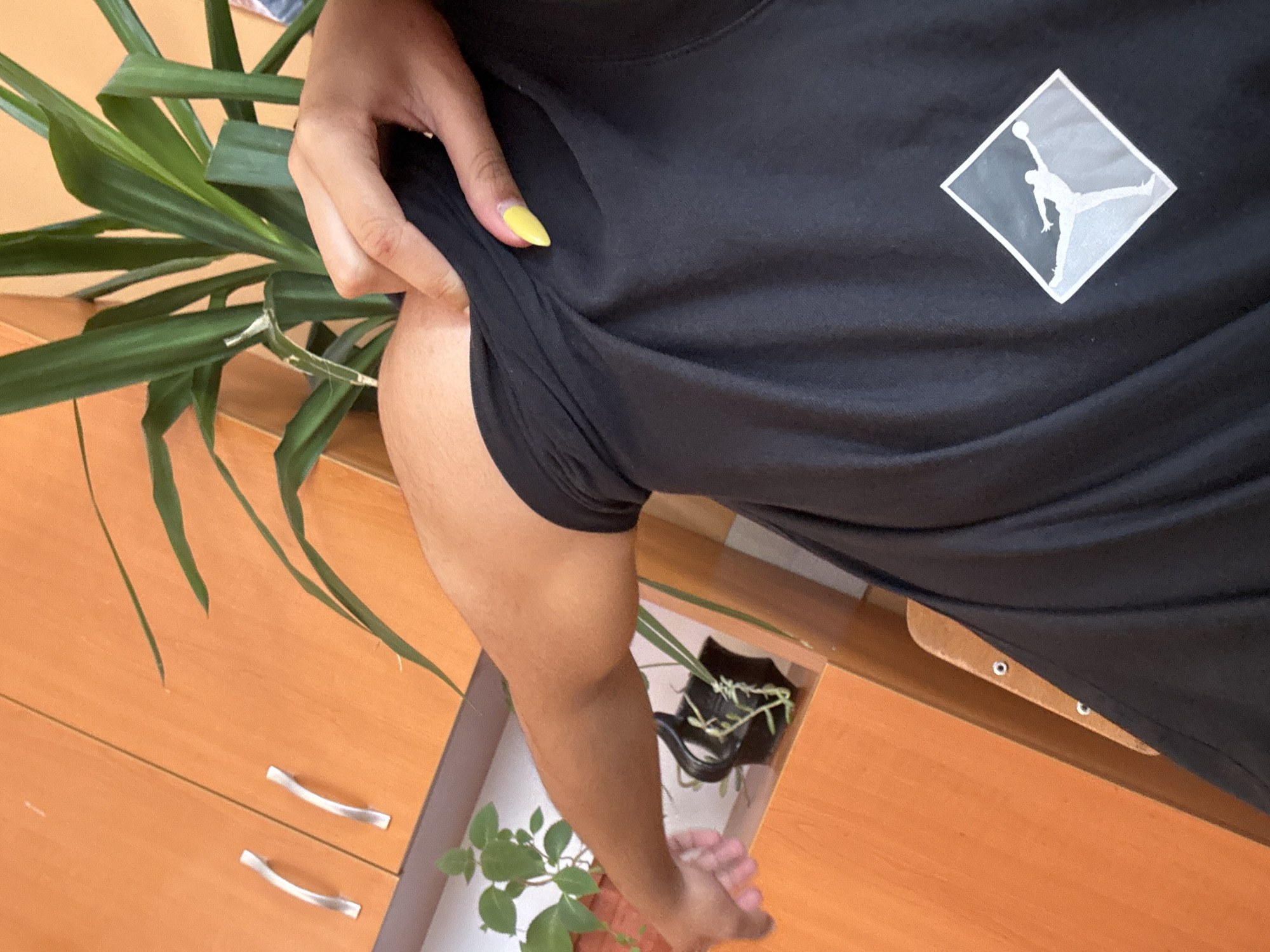 The image shows a person wearing a black t-shirt with a white Air Jordan logo standing next to a wooden dresser. The person has a yellow fingernail. A plant is visible on top of the dresser and in an open drawer below. The image was likely taken indoors. No information regarding the city of Buhovo, Bulgaria is provided.