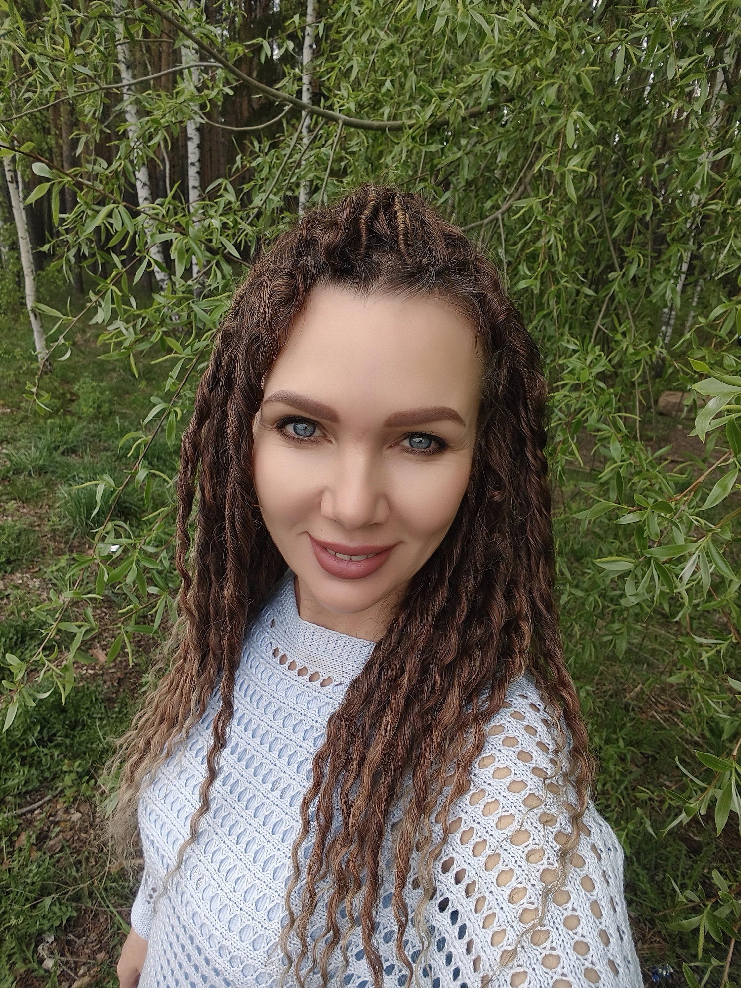 A woman with light skin and blue eyes smiles directly at the camera. Her hair is long, braided, and brown with lighter ends. She is wearing a light blue, open-knit sweater. The background consists of green foliage and birch trees. The location is outdoors, possibly in a park or wooded area. The city is unknown, unknown.