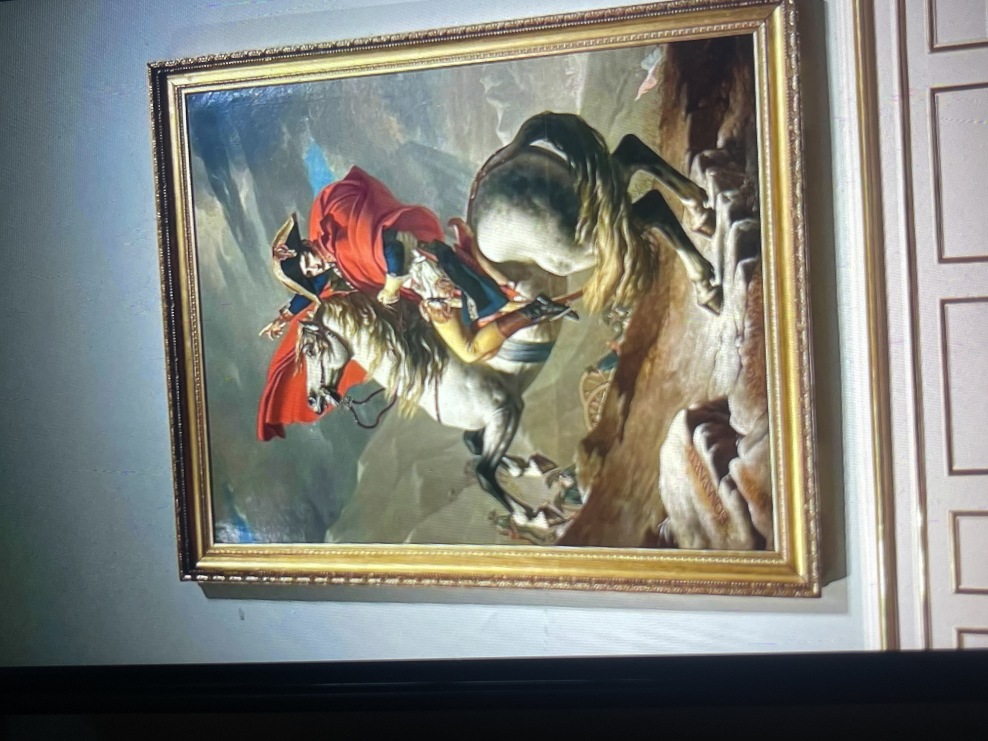 The image displays a framed painting of a man on a white horse. The man is wearing a red cape and a bicorne hat, and he is pointing with his left hand. The horse is rearing up on its hind legs. The background of the painting is a stormy sky and rocky terrain. The painting is hanging on a wall in what appears to be a museum or gallery, as indicated by the paneled wall below. The location is identified as Bishkek City, Kyrgyzstan.