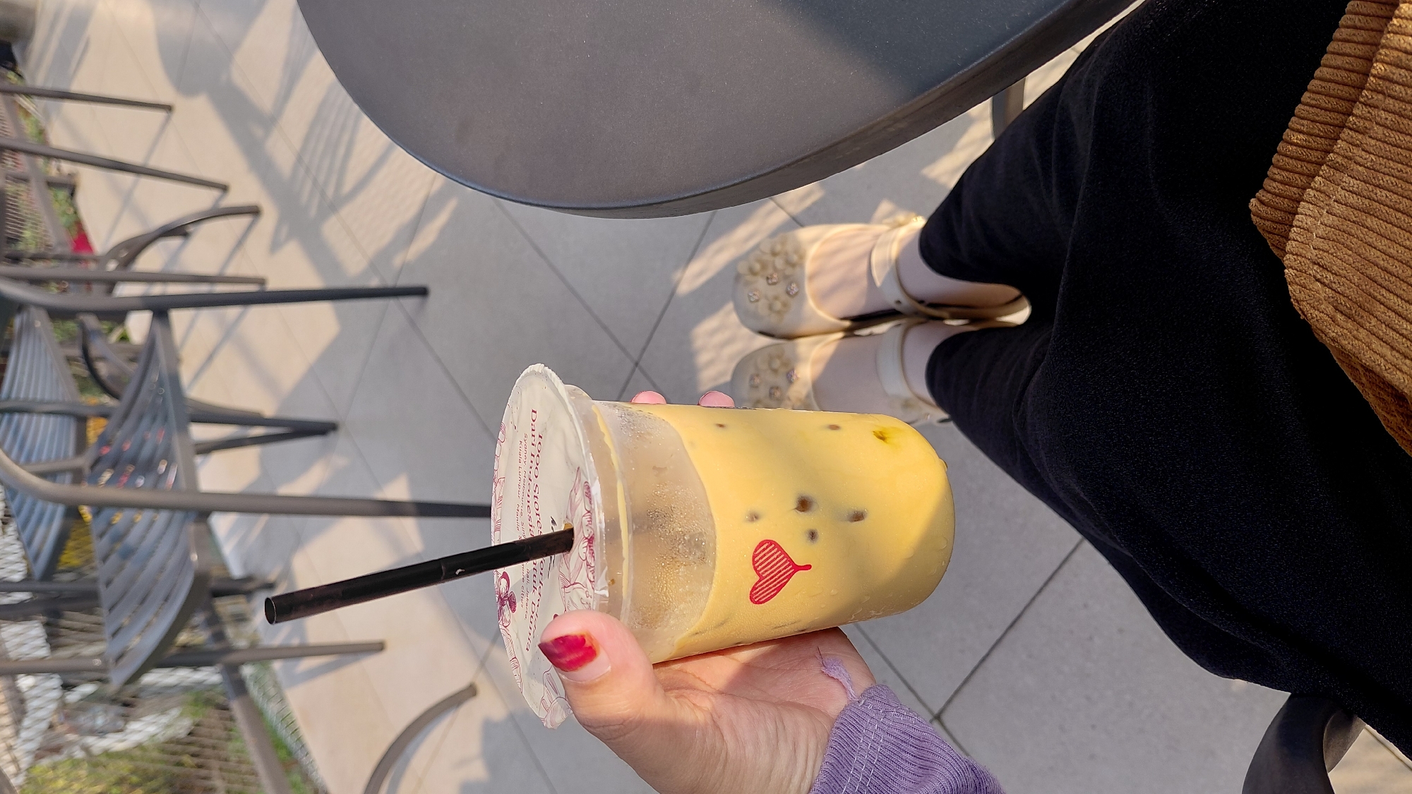 A person's hand, with red nail polish on the index finger, holds a clear plastic cup filled with a light yellow beverage containing ice cubes and a black straw. A small red heart icon is visible on the side of the cup. The person is wearing a purple long-sleeved garment and black pants. In the background, outdoor seating with metal chairs and tables is visible, suggesting a cafe or restaurant setting. The ground appears to be tiled. The scene is lit by natural sunlight, casting shadows from the chairs onto the ground. The presence of outdoor tables and chairs, along with the beverage, strongly indicates a public space like a cafe. The geometric tiling of the ground and the style of the chairs are common in urban cafes. Considering the prompt's request for a location in Karanglewas, Indonesia, this scene is consistent with an outdoor seating area of a cafe in that region.