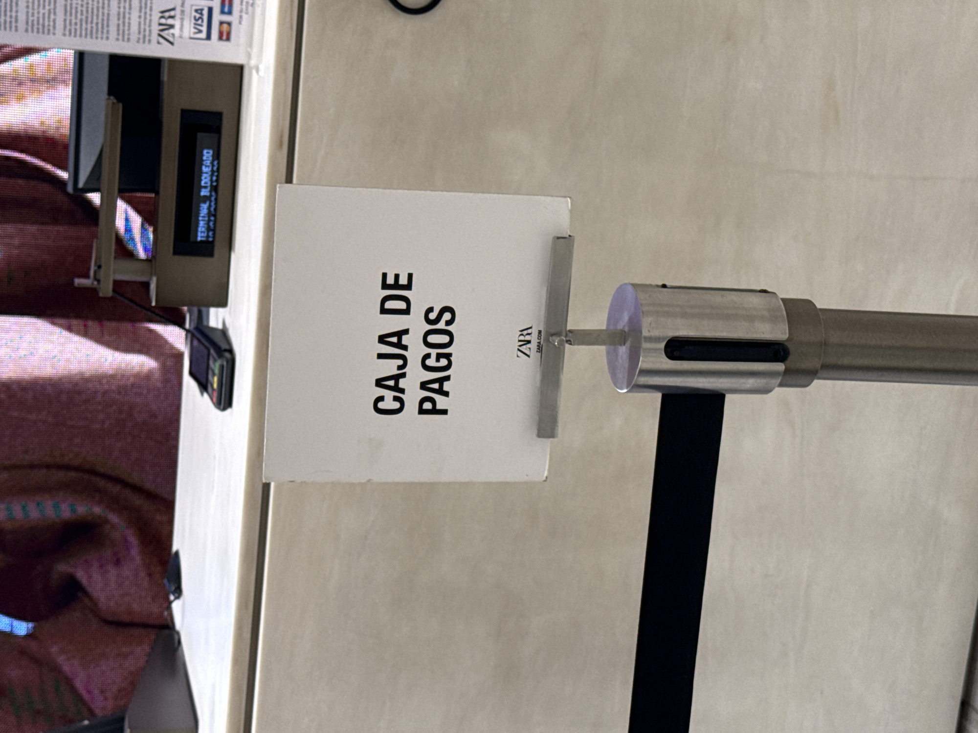 The image depicts a stanchion barrier with a sign reading "CAJA DE PAGOS" (Cashier) at a Zara store. The location is likely in the city of Puebla, Mexico, based on the presence of the Zara logo on the sign and the terminal displaying "TERMINAL BLOQUEADO". A credit card terminal is visible in the background, indicating a point-of-sale location.