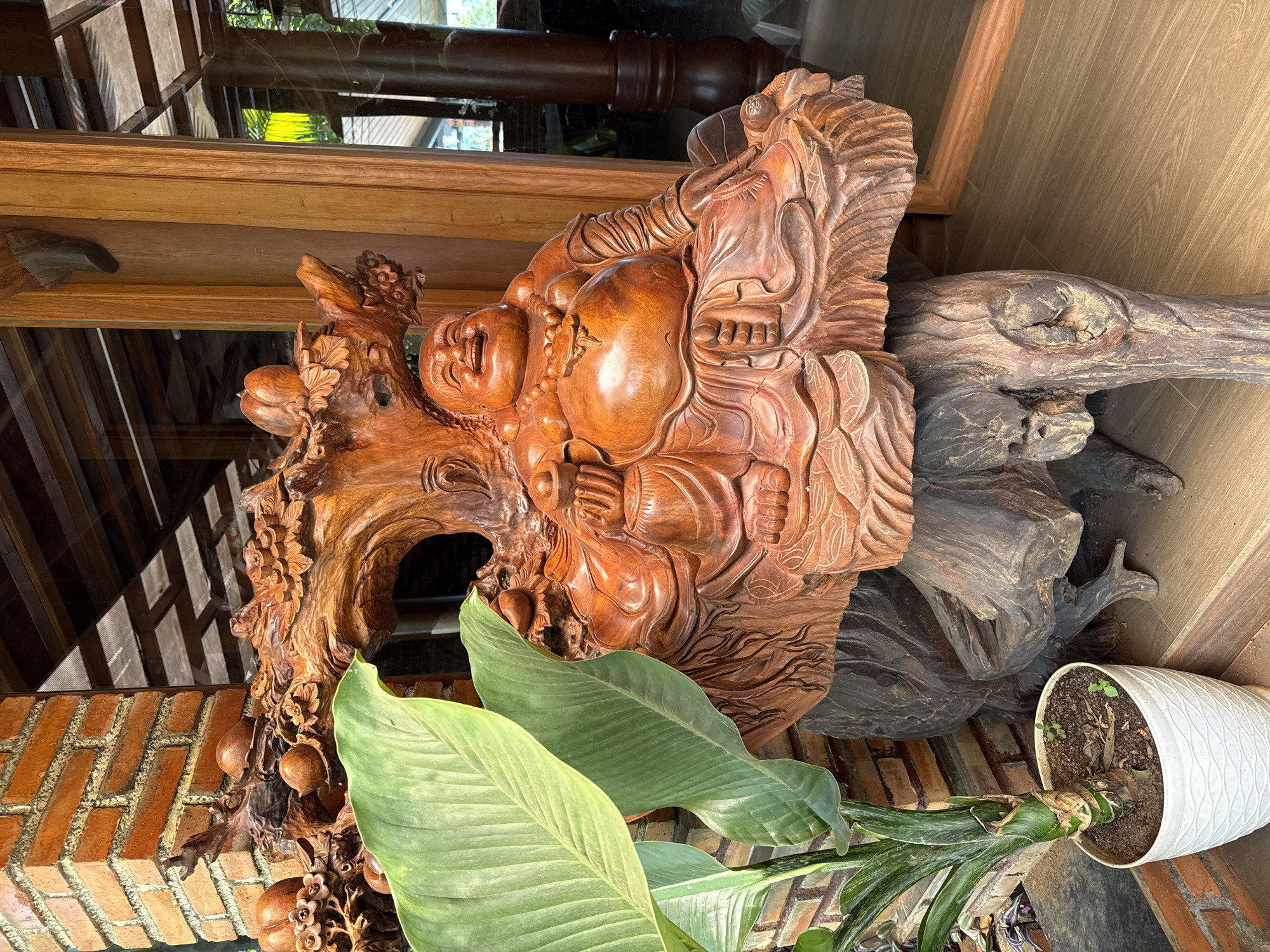 A carved wooden sculpture of a smiling Buddha figure is placed on a brick surface next to a potted plant. The sculpture is intricate, with detailed carvings of the Buddha and fruit-like decorations. The plant has large green leaves and sits in a white textured pot. The location is Chư Sê, Vietnam. The scene is indoors, with reflections of the surrounding environment visible in the glass behind the sculpture. The wooden sculpture is on a darker piece of wood.