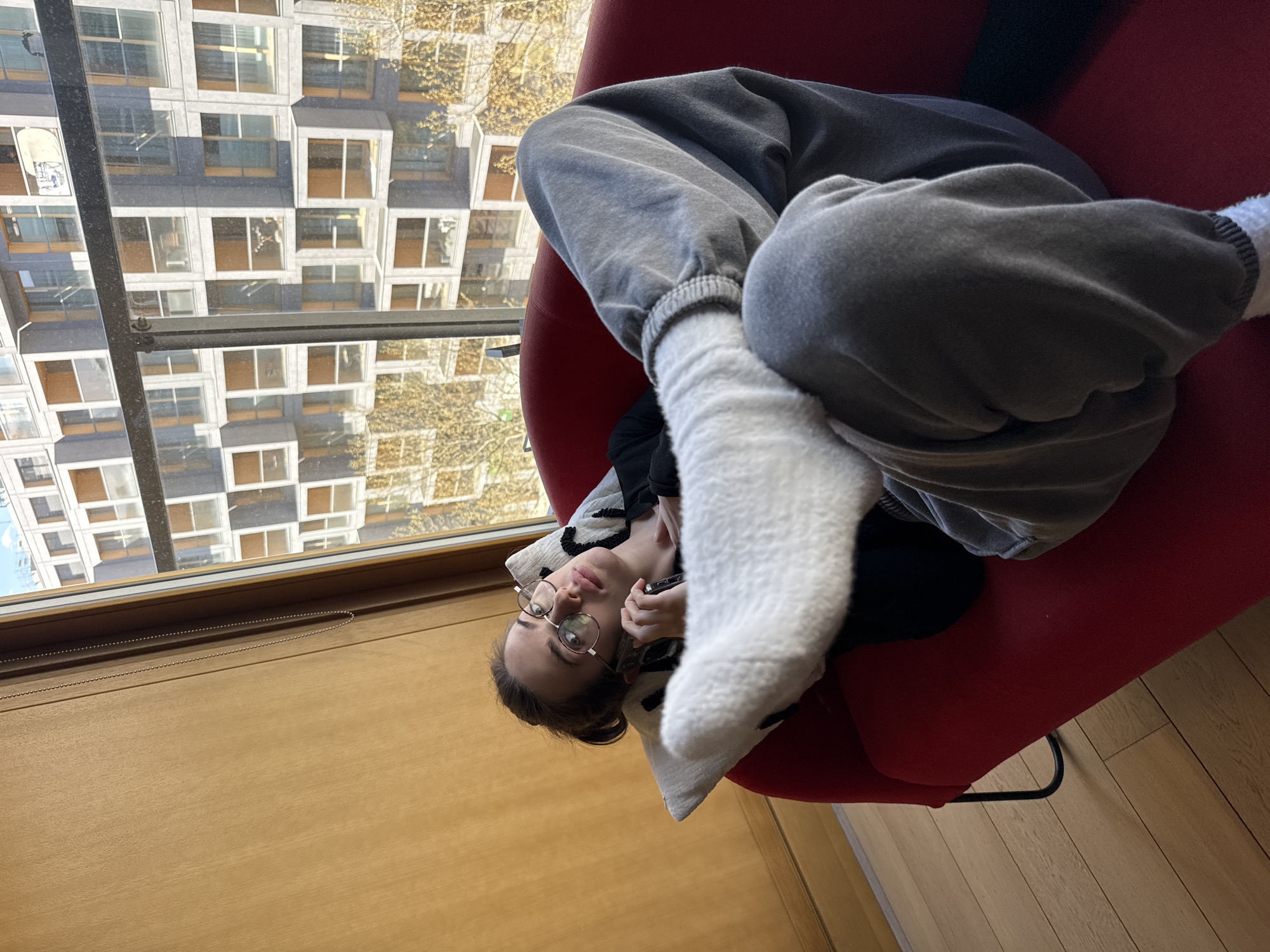 A young person with dark hair and round glasses is shown relaxing on a red armchair, positioned beside a large window. They are wearing grey sweatpants and white, fluffy socks, with their legs bent and feet raised. They appear to be lying on their back, looking towards the camera with a neutral expression, and are holding a dark-colored smartphone in their hand.
The interior features a light wooden floor and a wooden window sill, along which a thin beaded chain, possibly for blinds, is visible. Outside the window, a distinctive multi-story building with a facade composed of numerous square or rectangular windows and light-colored concrete frames is prominent. Beyond this building, trees with yellowish-brown autumn leaves suggest the season. The window also has dark metal framing, including a horizontal bar cutting across the view. The scene is brightly lit, indicating daytime. The overall image is rotated 90 degrees clockwise, creating an unusual perspective of the person in the chair and the outside view. The setting is an indoor space with a view of an urban environment, likely in Warsaw, Poland. No visible text is present.
