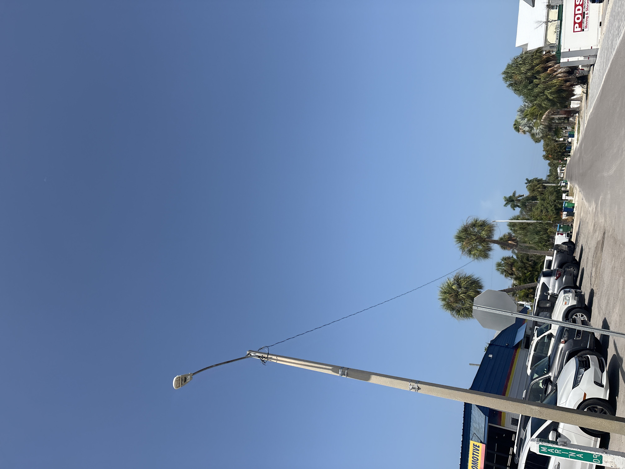 The image depicts a street scene in Holmes Beach, Florida, United States of America, on a clear, sunny day with a bright blue sky. In the foreground, a concrete utility pole stands to the left, supporting electrical wires. Adjacent to the pole is a white car with a green and white sign that reads "MARINA DR". Behind the car, a line of parked vehicles, including SUVs and cars, are visible. To the left, a building with a blue facade and yellow lettering that reads "AUTOMOTIVE" is partially visible. Further down the street, palm trees and other vegetation line both sides. In the distance, the street recedes towards a row of buildings. On the right side of the street, a white PODS container with "Moving & Storage" branding is parked. The environment appears to be a commercial or mixed-use area, possibly near a coastal location, given the presence of palm trees and the name of the street. There are no people clearly visible in the image.