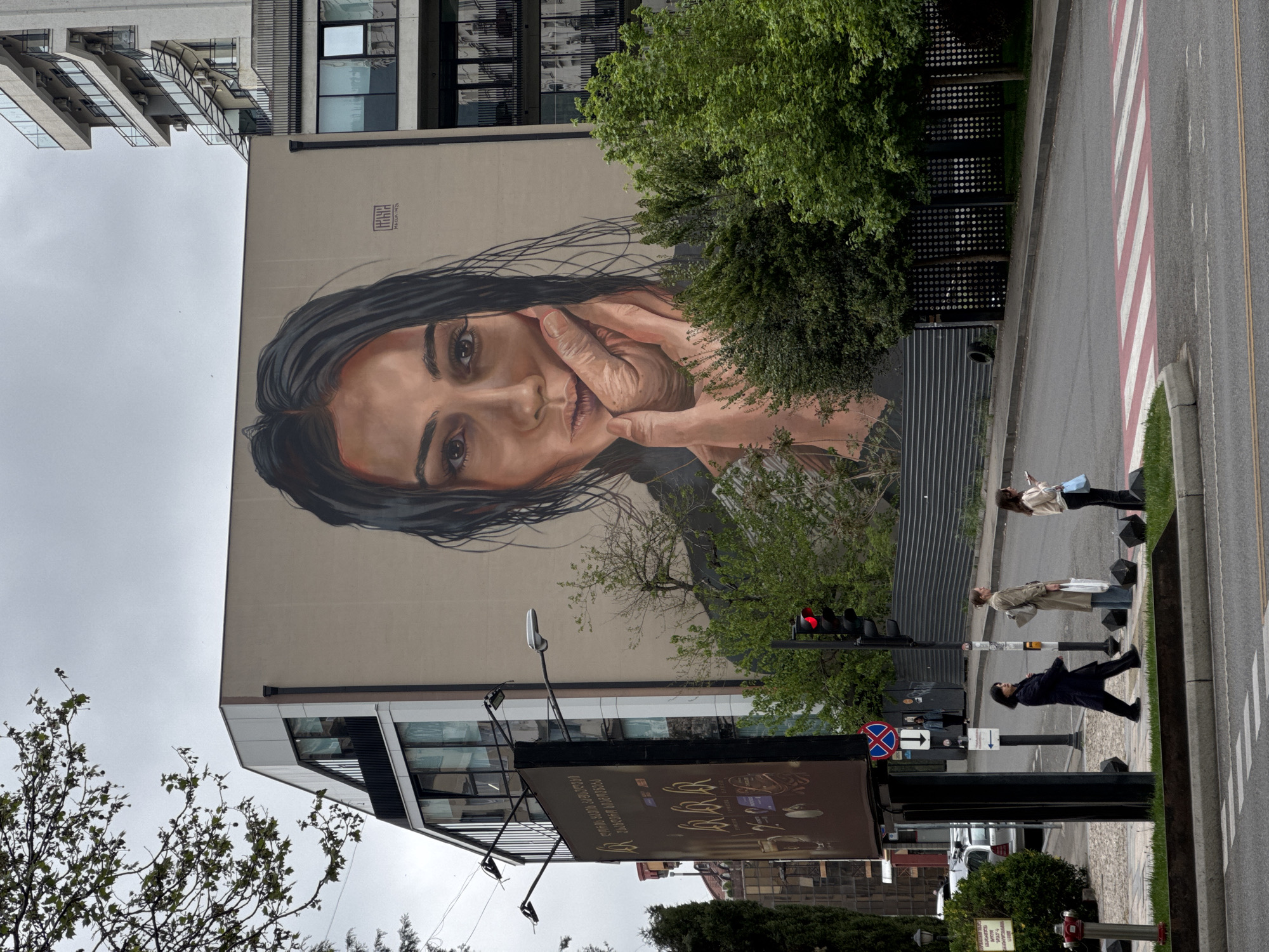A large mural of a woman's face is painted on the side of a building in Tbilisi, Georgia. The woman has dark hair and is looking directly at the viewer with her hands near her face. In the foreground, there is a billboard advertising a product, a traffic light showing red, and a crosswalk with pedestrians waiting to cross the street. The sky is overcast, and there are buildings and trees visible in the background.
