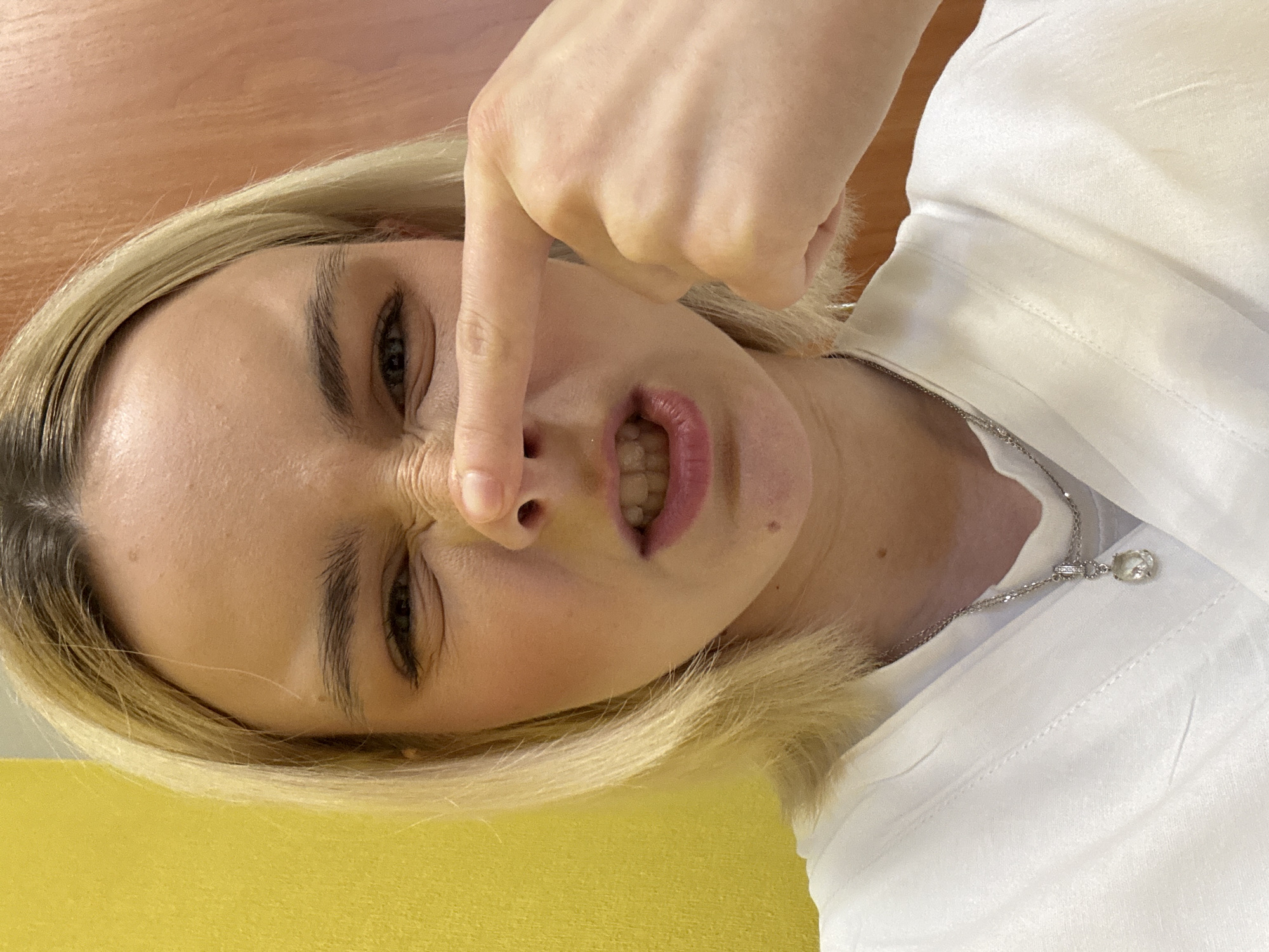 A woman with blonde hair is holding her nose with her index finger. She is wearing a white shirt and a silver necklace. The background is a yellow surface. The image does not depict any information to imply its relation to Saint Petersburg, Russia.