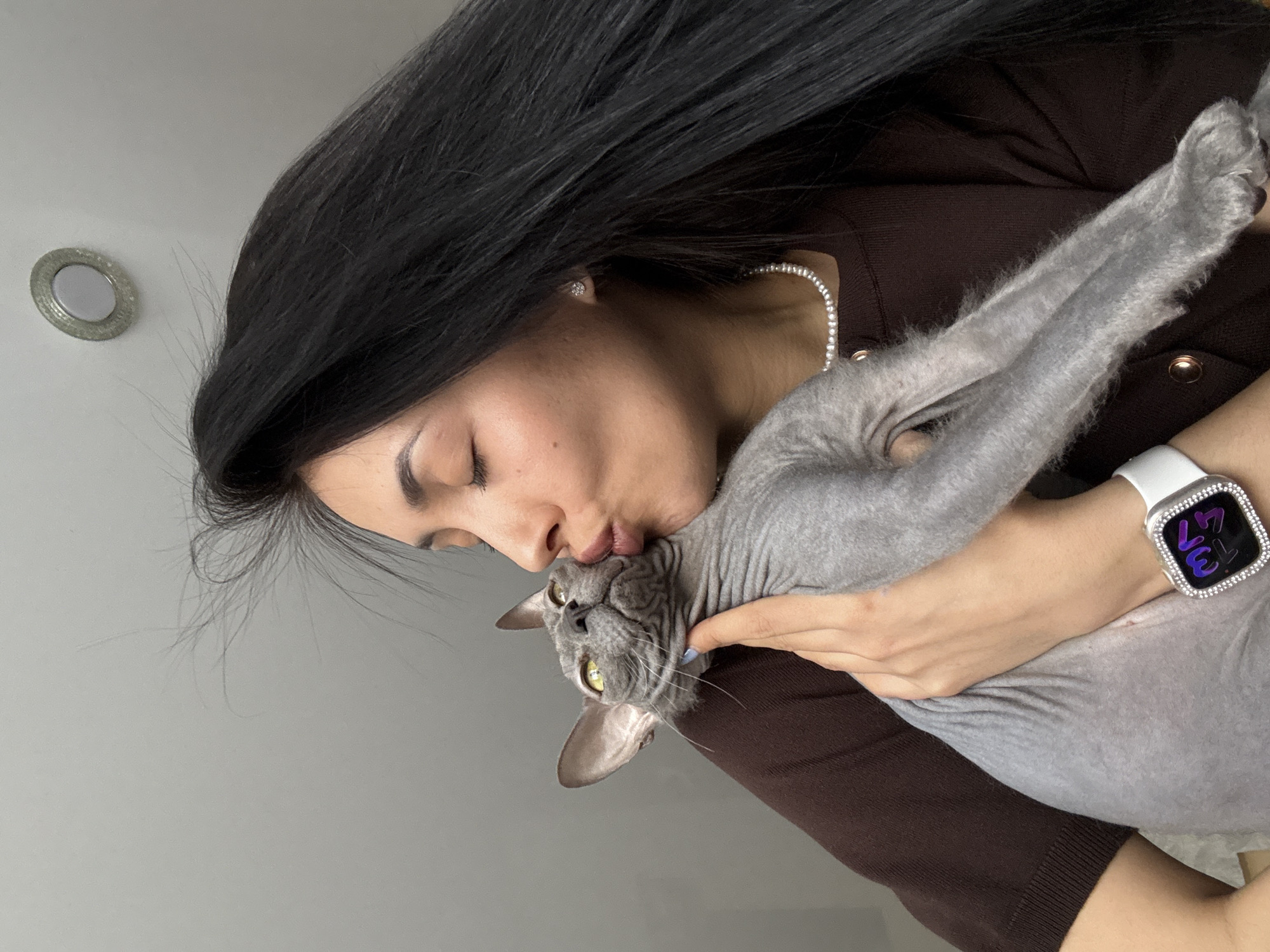 A woman with long, dark hair is kissing a gray hairless cat. She is wearing a brown long-sleeved shirt and a white smartwatch. The background is a plain gray wall. There is a light fixture on the wall above her head. The location is not discernable from this image and therefore cannot be narrowed down to Votkinsk, Russia.