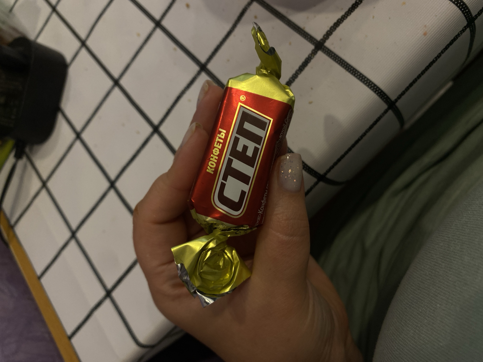 A hand holds a red and gold wrapped candy with the Russian word "СТЁП" in large white letters with a red outline. The word "КОНФЕТЫ" appears above "СТЁП". The candy is held over a white surface with a black grid pattern. In the background, out of focus, is a black electrical device and a purple object. The scene is likely indoors and does not provide any specific location details within the city of Lomonosov, Russia.