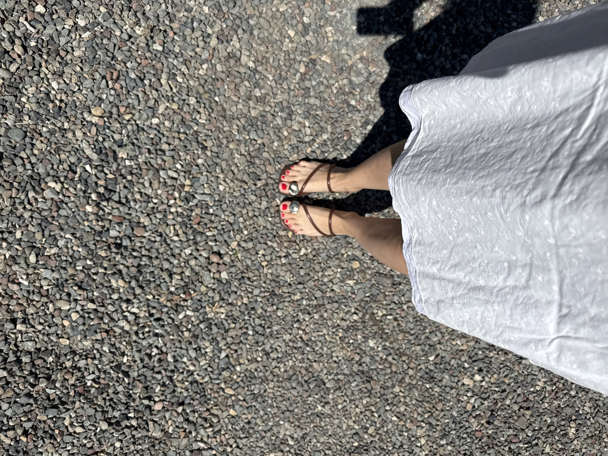 A top-down, medium shot shows a person's bare feet and the lower part of their white dress on a gravel surface. The feet are adorned with bright red nail polish and brown sandals with circular embellishments. A shadow of the person is cast on the gravel to the right. The background consists of a dense scattering of grey and brown pebbles. The scene appears to be outdoors.