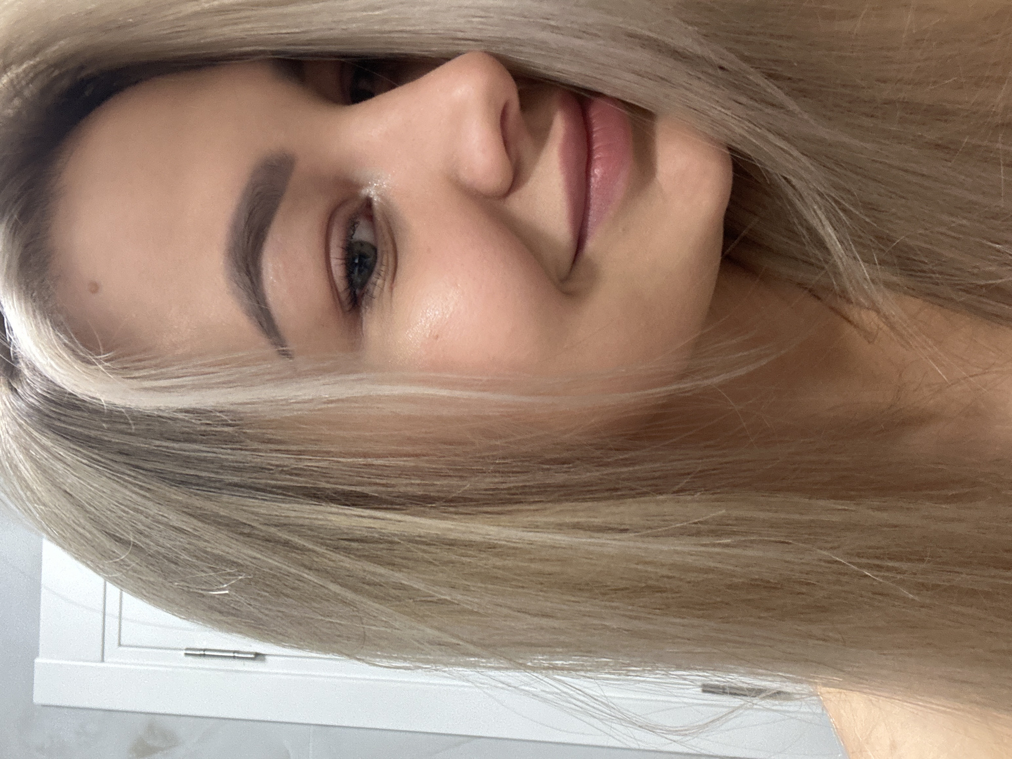 A close-up portrait captures a woman with light blonde hair. Her hair partially obscures her face. Her facial features include an eyebrow, an eye, a nose, and lips. The background appears to be a light-colored wall. The location of the image cannot be determined from this image alone.