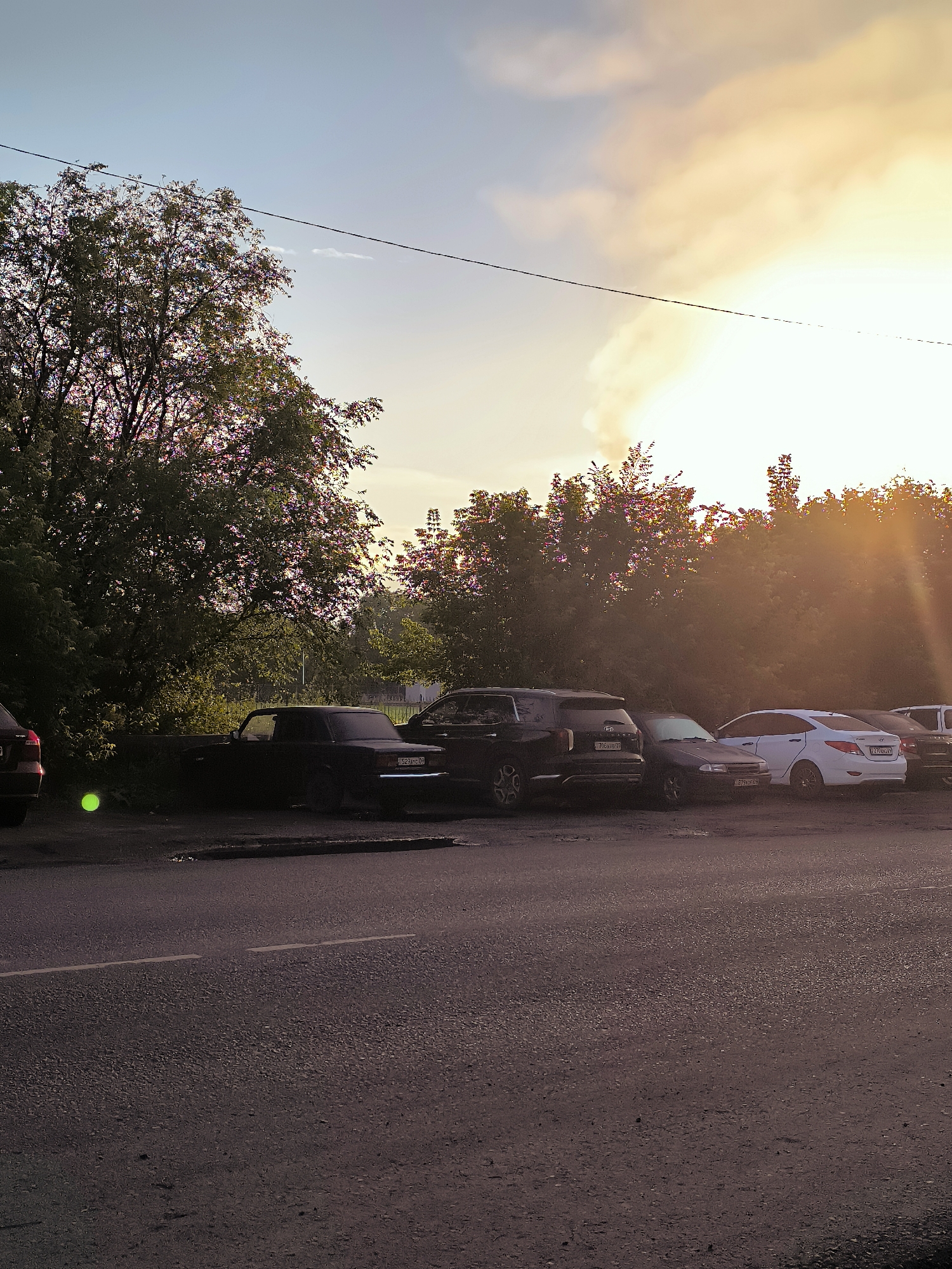 The image depicts a street scene in Qarağandı, Kazakhstan, at dusk or dawn, with warm, golden light filtering through the trees and clouds. A line of parked cars is visible along the side of a paved road. The cars appear to be a mix of older and newer models, including a dark-colored sedan, an SUV, and a white sedan. Trees with lush green foliage line the background, with some trees exhibiting red and pink hues, possibly due to flowering or the setting sun. The sky is a gradient of pale blue to warm orange and yellow, with some wispy clouds. A power line is stretched across the upper part of the frame. The road in the foreground has a white line marking. The overall impression is a quiet, everyday scene in a residential or commercial area of the city.