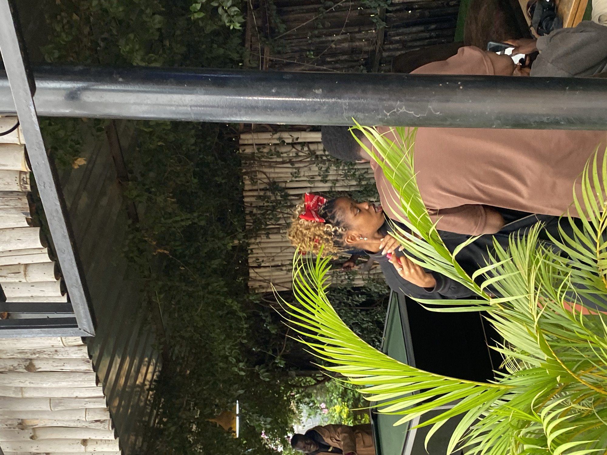 In Gura Top, Nigeria, amidst lush foliage and bamboo accents, a person with curly blonde hair adorned with a red bow is captured in mid-action, holding a pink object and looking down. To their right, another individual in a brown t-shirt faces away from the camera, holding a phone. In the background, a person in a brown jacket smiles. The setting appears to be an outdoor venue with a dark green pool table visible. A prominent black pole stands in the foreground, partially obscuring the view.