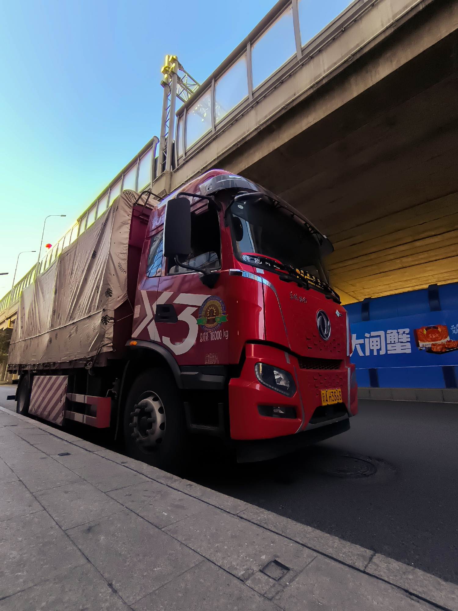 A red flatbed truck with a covered cargo bed is parked on the right side of a city street, partially under a concrete overpass. The truck has the logo "X3" in large white lettering on its side, along with some text and a circular emblem. The truck's license plate, "A H3868", is visible. To the right of the truck, a blue barrier with Chinese characters and an image of food is present. The street appears to be in an urban environment with sidewalks and streetlights. The sky is clear and blue. The overall scene is a snapshot of a vehicle parked in a city, likely in Ürümqi, China, as indicated by the presence of Chinese text on the barrier and the vehicle.