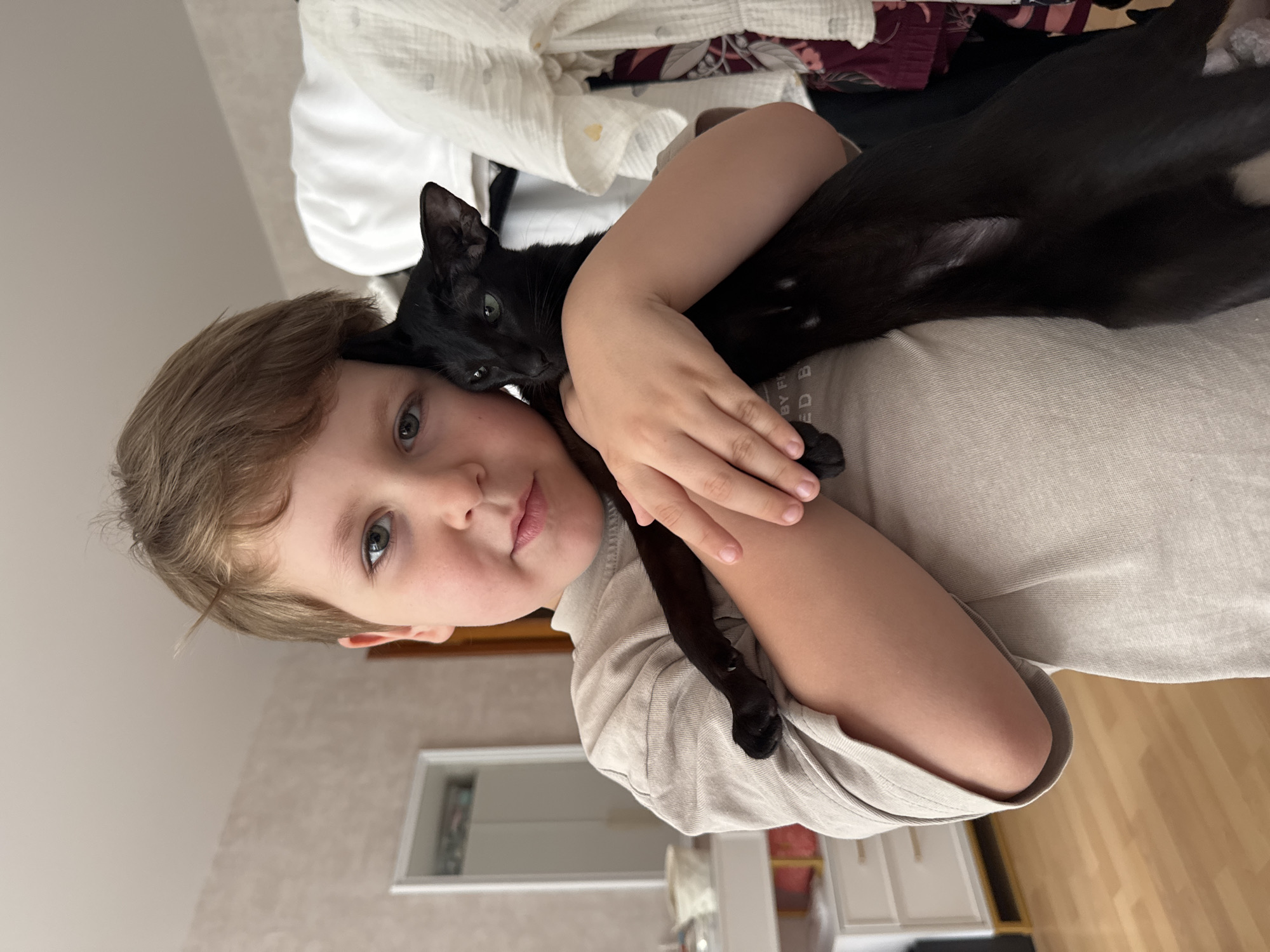A young boy with light brown hair is holding a black cat. The boy is wearing a beige t-shirt. He is looking at the camera with a slight smile. The cat is nestled in his arms and is looking at the camera with green eyes. The background appears to be a bedroom with a dresser and a closet visible. The lighting suggests it is daytime.