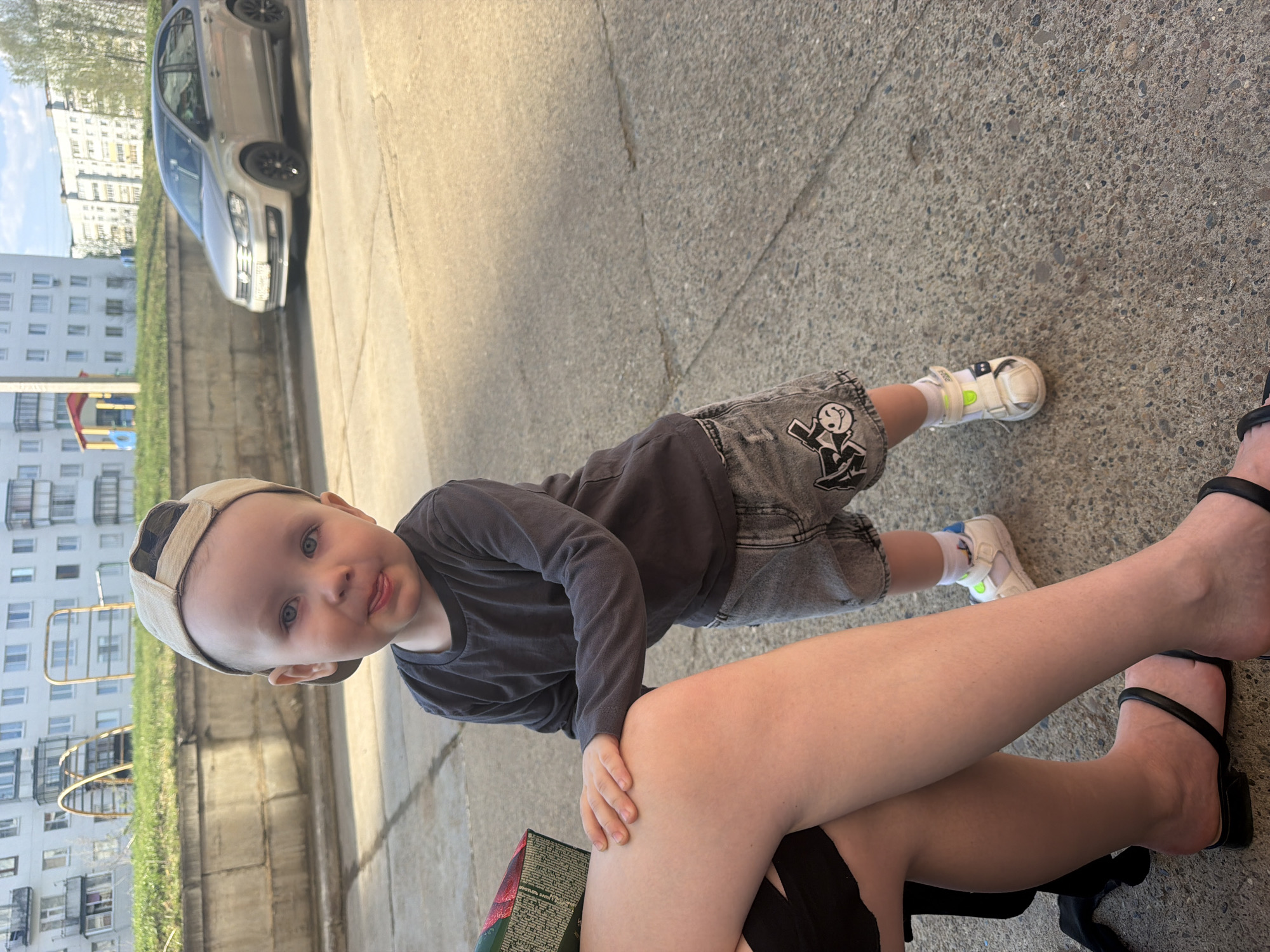 A toddler is standing on a sidewalk, leaning on the knee of a person seated in a chair. The toddler is wearing a tan baseball cap, a dark gray long-sleeved shirt, gray denim shorts with a cartoon graphic, and white shoes with blue and lime green accents. The seated person is wearing black sandals with a block heel and a black garment. A portion of a food container with red and green packaging is visible near the person's knee. In the background, a silver car is parked along the curb. Across the street, a tall apartment building with rows of windows and metal balconies is visible, as is a playground with a metal climbing structure and green grass. The photograph was taken during the daytime, with bright sunlight casting shadows. The location appears to be a residential area in the city of Chaikovsky, Russia.
