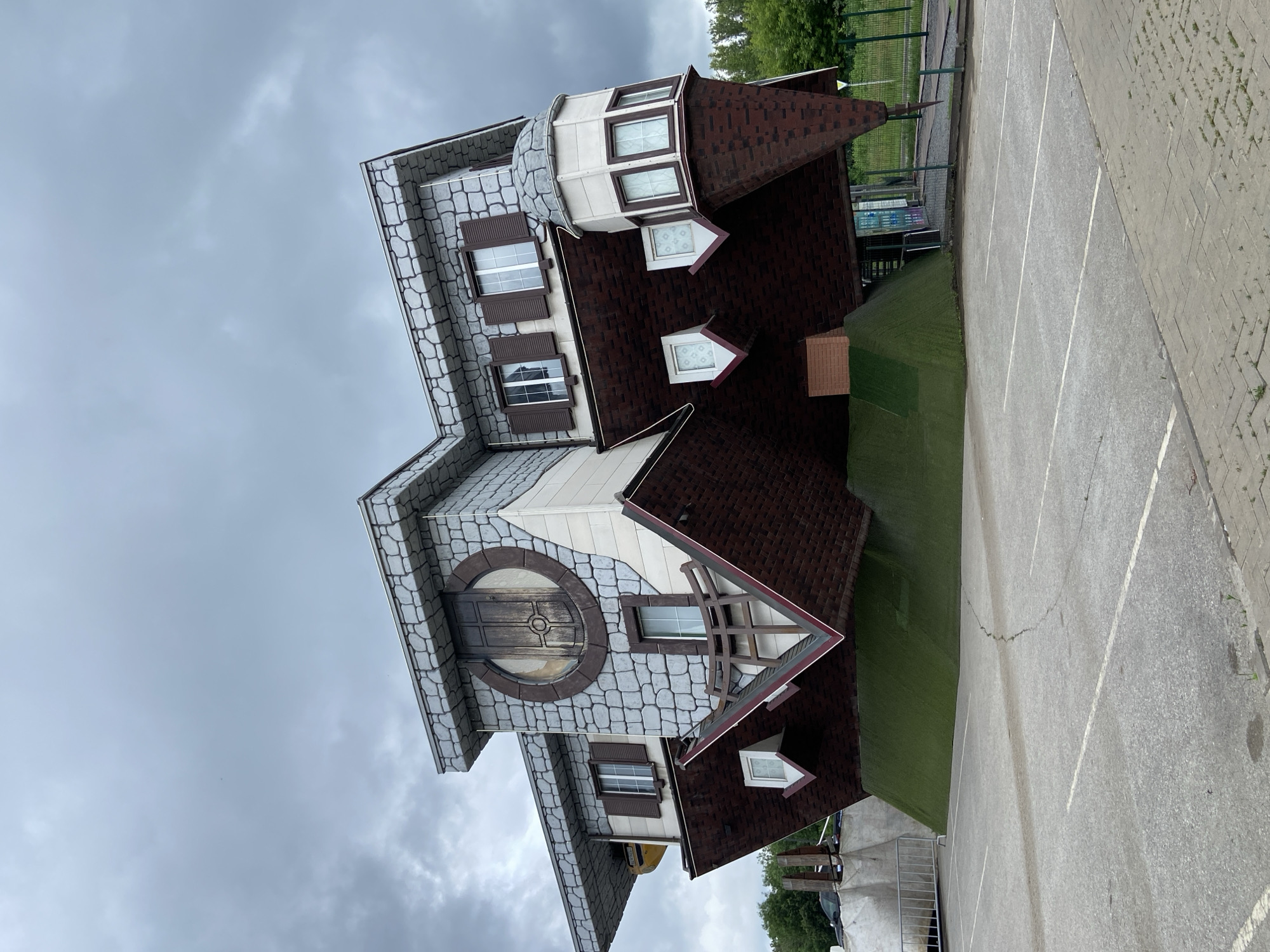An upside-down house, designed to appear as if it's been flipped over, is situated in a paved parking lot in Ufa, Russia. The structure features a grey stone-patterned facade on its upper section, with several windows, some adorned with brown shutters. A large, round wooden door with intricate carvings is centrally located on the upper part of the house. The lower section of the house is covered in dark red, shingle-like material, mimicking a roof, and extends downwards, creating the illusion of being the foundation. Small, white-framed windows with patterned glass are visible on this inverted roof portion. A green, artificial turf-covered slope forms a base for the upside-down house, extending into the parking lot. The sky above is overcast with grey clouds. In the background, a metal fence separates the property from a grassy area and trees. The parking lot has visible white painted lines marking parking spaces.