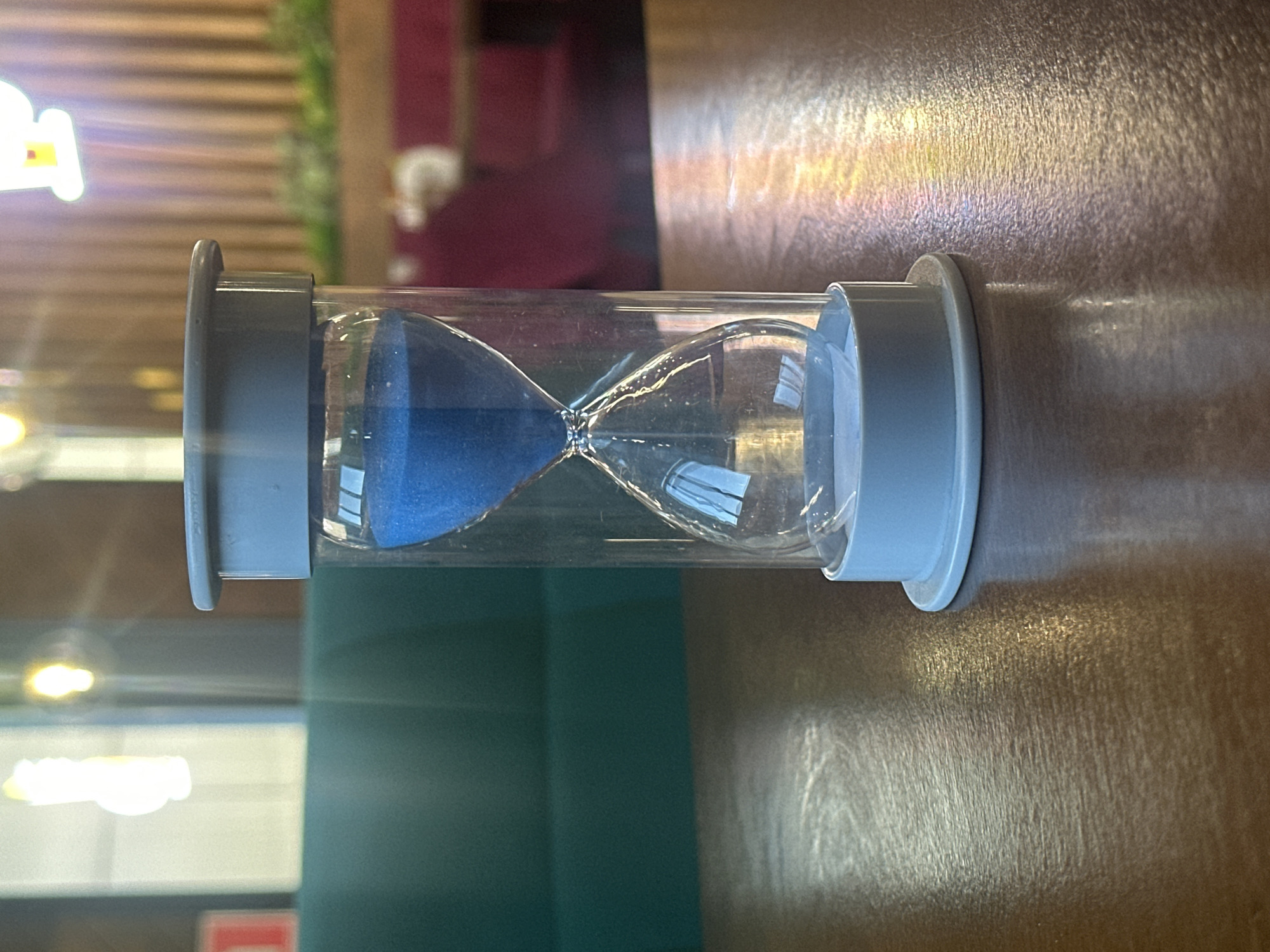 An hourglass with blue sand sits on a wooden table. The hourglass is made of glass with a light blue plastic frame at the top and bottom. The sand is mostly in the top bulb, with a small amount having fallen into the bottom bulb, indicating that time has begun to pass. The table surface is a medium-dark wood with a visible grain and reflects light. In the background, out of focus, is a dimly lit indoor setting that appears to be a restaurant or cafe. A teal-colored couch is visible, along with some wooden paneling on a wall, and soft lighting fixtures. There are no people or animals present in the image, and no actions or interactions are depicted. The location is identified as Blagoveshchensk, Russia, based on external information, but this cannot be determined from the visual content of the image itself.