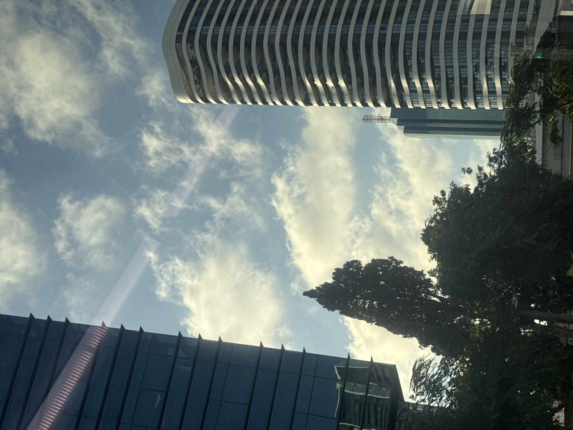 The image captures a view looking upwards towards a bright sky filled with scattered white clouds, suggesting a clear day. The sunlight creates lens flares, indicating the photo was taken with a camera or phone. In the foreground, a modern building with a dark, reflective facade and a sharp, angular top is visible. The glass panels of this building reflect the sky and surrounding structures.
Dominating the upper portion of the image is a large, curved skyscraper with a distinctive facade composed of horizontal white and grey bands, creating a wave-like appearance. The building has many windows, suggesting office spaces or apartments. Further in the background, another taller, rectangular skyscraper with a blue-green glass exterior is visible. A cellular tower can be seen on top of this building.
To the right of the frame, several large, dark green trees with dense foliage partially obscure the view of the buildings and sky. One particularly tall, slender tree stands out. The overall scene depicts an urban environment, likely a cityscape, given the presence of multiple tall buildings. The orientation of the image, with the sky above the buildings, suggests the photo may have been taken from the ground looking up, or potentially from within another building. The lighting suggests it is daytime, likely morning or afternoon. There are no people or specific events depicted, focusing instead on the architecture and natural elements within an urban setting. The location is identified as Tel-Aviv, Israel.