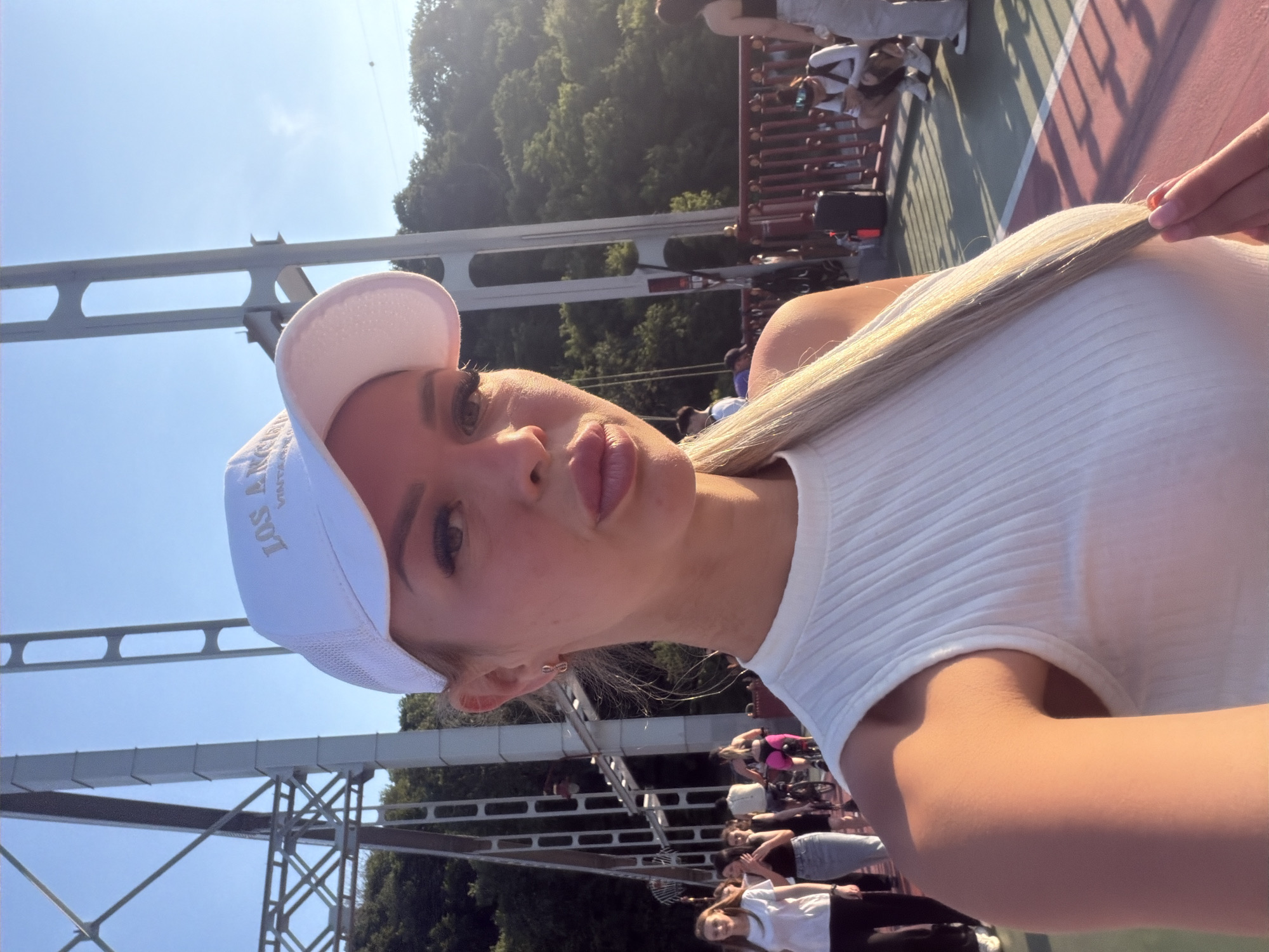A blonde woman wearing a white visor cap and a white ribbed tank top takes a selfie on a pedestrian bridge in Kyiv, Ukraine. The woman's face is close to the camera, with her mouth slightly parted. Her blonde hair is pulled back into a ponytail, and she is holding a strand of it with her right hand. In the background, there are other people walking and biking on the bridge, as well as trees and a clear blue sky. The bridge appears to be a modern structure with a steel framework. The lighting suggests it is late afternoon.