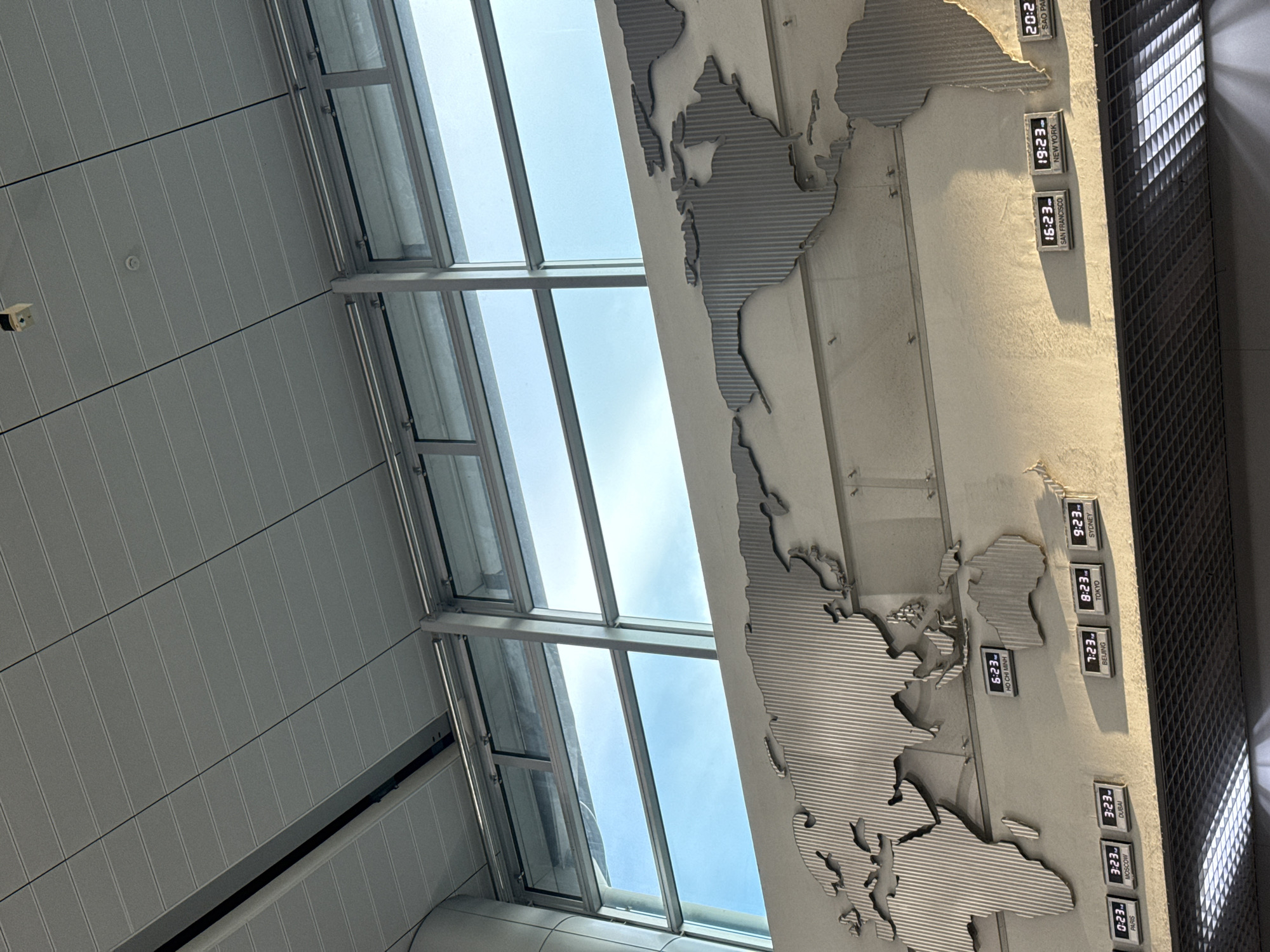 This image shows a world map decorated on a wall, with digital clocks displaying different city times. The map appears to be made of metal and has a textured surface. Below the map, several digital clocks are visible, showing times for cities such as Paris, Moscow, Dubai, Ho Chi Minh, Beijing, Tokyo, Sydney, San Francisco, New York, and Sao Paulo. The clocks are illuminated and appear to be functioning. The background consists of a large window with a bright sky visible through it, suggesting an indoor location with natural light. The ceiling above appears to be made of modular panels. The overall scene depicts a modern interior space, possibly an airport or a public building, with a focus on global time representation. While the question mentions Thuận An, Vietnam, there is no visual information within the image that directly identifies this specific location. However, the presence of a clock for Ho Chi Minh City might suggest a connection to Vietnam.