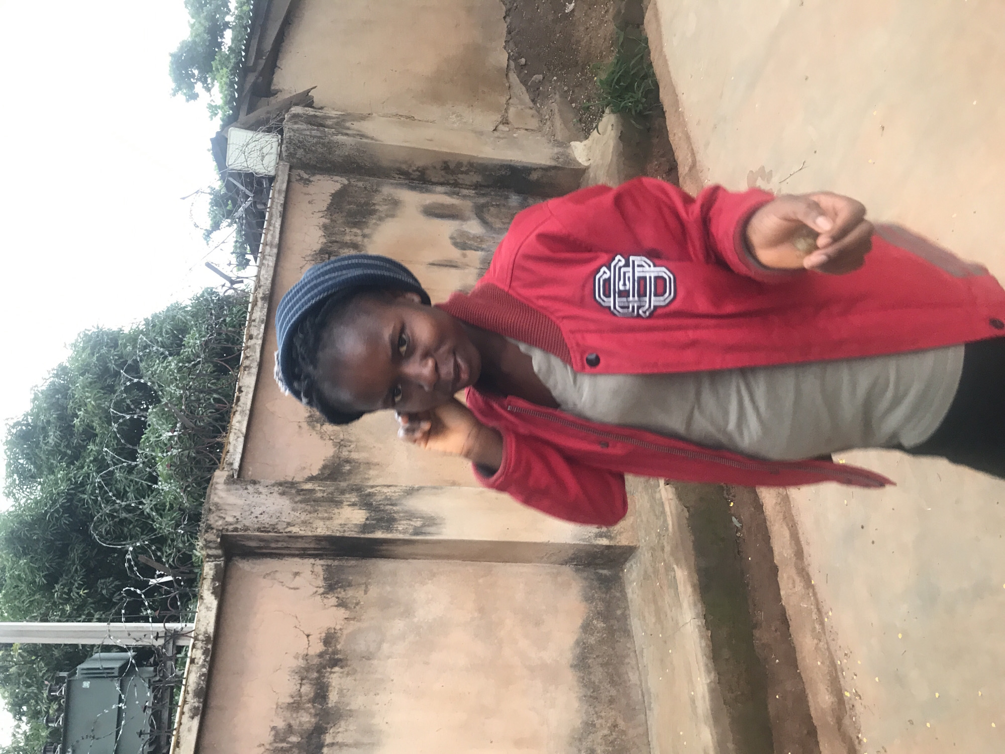 In Kabong, Nigeria, a young Black woman stands against a weathered, pale peach-colored wall. She is wearing a red bomber jacket with a white and black logo on the left breast, over a grey t-shirt. A blue and white striped beanie covers her hair. Her right hand is raised to her ear, and her left hand is extended, holding a small object. The background features a metal fence topped with barbed wire, trees, and a large electrical transformer. The ground is a sandy, earthen path.