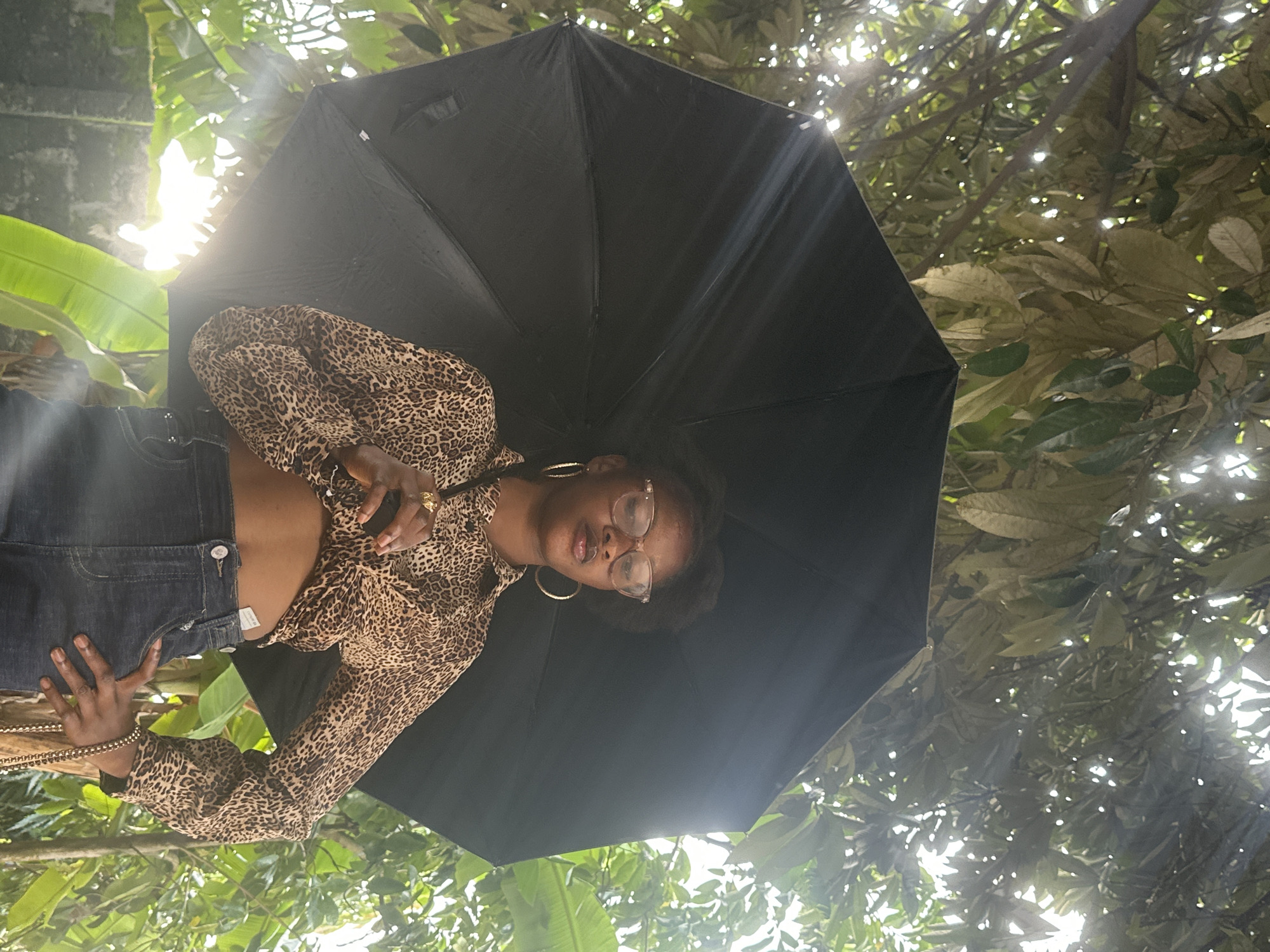A low-angle, eye-level, medium shot features a young Black woman standing outdoors in an environment that suggests the city of Badagry, Nigeria, with abundant green foliage and bright sunlight filtering through the trees. She holds open a large black umbrella, which partially obscures her. The woman wears a long-sleeved, leopard-print, cropped shirt and dark denim jeans. She has on clear-rimmed glasses, large hoop earrings, and several rings on her fingers. Her left hand rests on her hip, and her right hand holds the umbrella handle. The background is filled with lush green leaves and banana plants, with a hint of a stone wall visible to the left. The lighting suggests a sunny day, with lens flare visible due to the bright light.