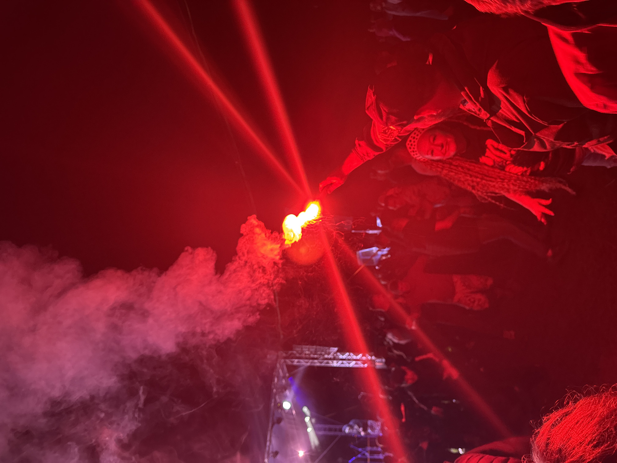 The image depicts an outdoor night event in Большая Кивара, Russia, characterized by a dominant red light spectrum. A large crowd of individuals occupies the lower and mid-ground, largely appearing as dark silhouettes due to the low light and heavy red illumination. In the upper-middle right, a person's arm and hand are visible holding a lit red flare or smoke bomb, which emits bright orange-red flames and dense red smoke plumes that ascend towards the upper left. This flare is a significant source of the red light and smoke in the central area. Multiple parallel red laser beams cut across the upper right portion of the scene.
On the far left, a stage structure constructed from metal scaffolding is visible, equipped with several lights emitting white and blue hues, contrasting with the pervasive red. A performer or individual is discernible on the stage. In the lower right foreground, a person with long, braided hair, wearing a patterned headband and a dark shirt with a lighter graphic, is partially visible, illuminated by the red light. The overall environment is heavily impacted by the smoke and intense red lighting, creating a high-contrast, atmospheric scene. Attendees are dressed in casual, dark attire.