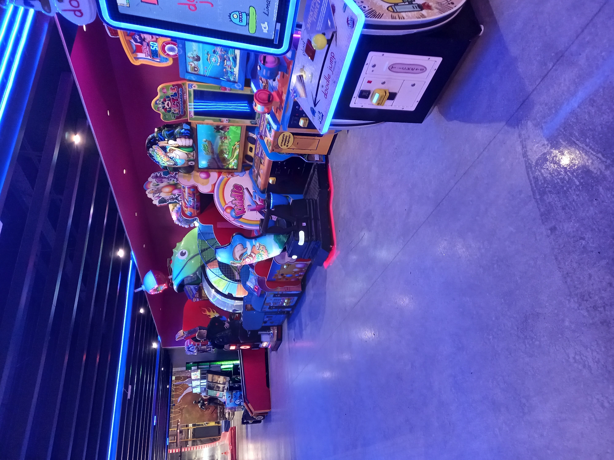 An indoor amusement arcade in Yekaterinburg, Russia, features rows of brightly lit arcade machines. The floor is concrete, and the ceiling has a dark, slatted design with blue neon lighting. The arcade machines display various games with colorful graphics and cartoon characters. Some machines have screens showing game animations, while others have control panels with joysticks and buttons. The machines are illuminated by their own lights and the ambient neon. A few people are visible in the background, one of whom is standing near some machines. The overall atmosphere is vibrant and exciting, typical of an entertainment venue.