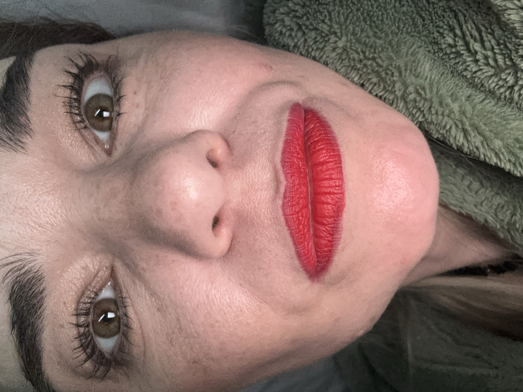 A close-up photograph captures a woman's face tilted to the left. She has fair skin, brown eyes, and dark eyebrows. Her lips are painted with a bright red lipstick, which accentuates the vertical lines on the lips. The background is a mix of gray and green, with a textured green fabric partially visible. No specific details point to a location in Murino, Russia; the image focuses solely on the subject's face and immediate surroundings.