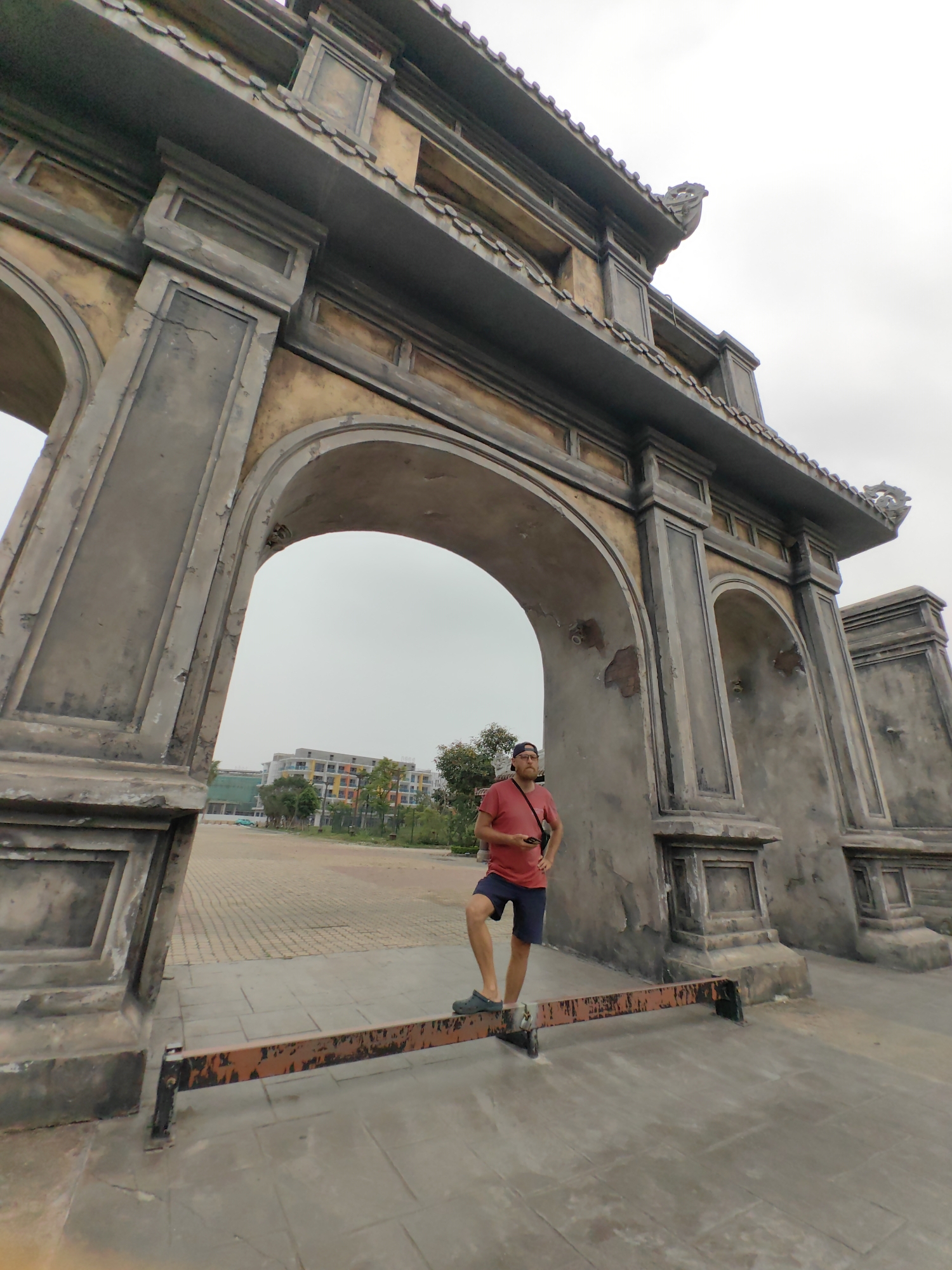 A low-angle, wide-shot depicts a man standing on an orange and black metal bar between two large, ornate gray and tan arches. The man, presumed to be Caucasian, is wearing a red t-shirt and dark blue shorts with gray shoes. He has a beard and is wearing a dark baseball cap and a camera on a strap across his chest.
The arches, of apparent Vietnamese design with stylized dragon ornamentation on the roof, are large and dominate the scene. The sky is overcast.
Behind the arches is a view of a building with colorful walls and several trees. The paving in front of the arches is gray stone. The location is Bai Chay Ward, Vietnam.