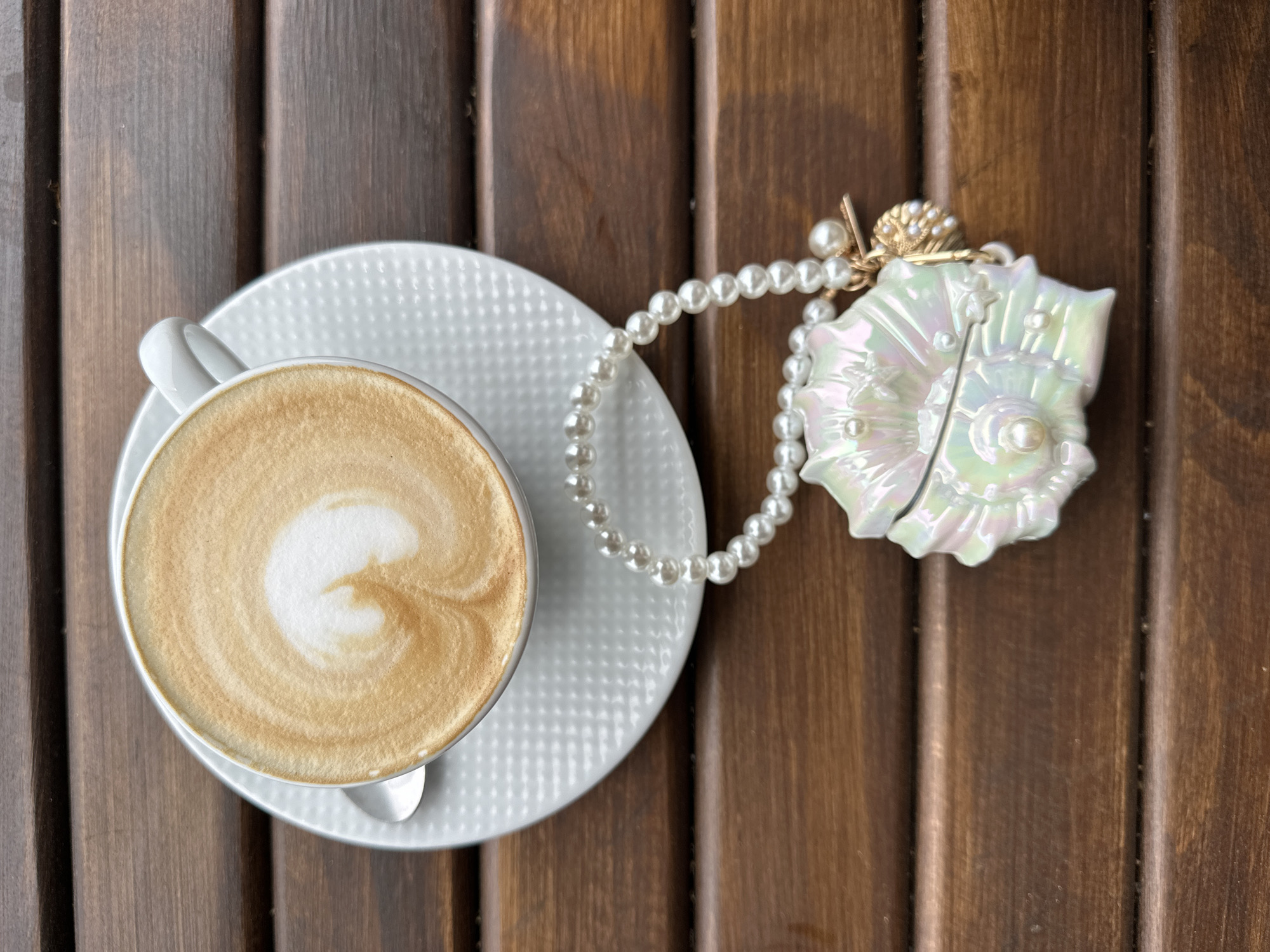 A top-down, eye-level, close-up shot on a dark brown, planked wooden surface features a cup of coffee on a patterned white saucer, adjacent to a strand of faux pearls and a shell-shaped brooch. The coffee, served in a white ceramic mug, has a latte art swirl on the surface. The strand of pearls gently curves towards the brooch, which has a pearlescent sheen. The brooch is mostly off-white in color. The photograph appears to have been taken in natural lighting. The location (Šušanj, Montenegro) is not evident from the image itself. No people, actions, or interactions are visible.