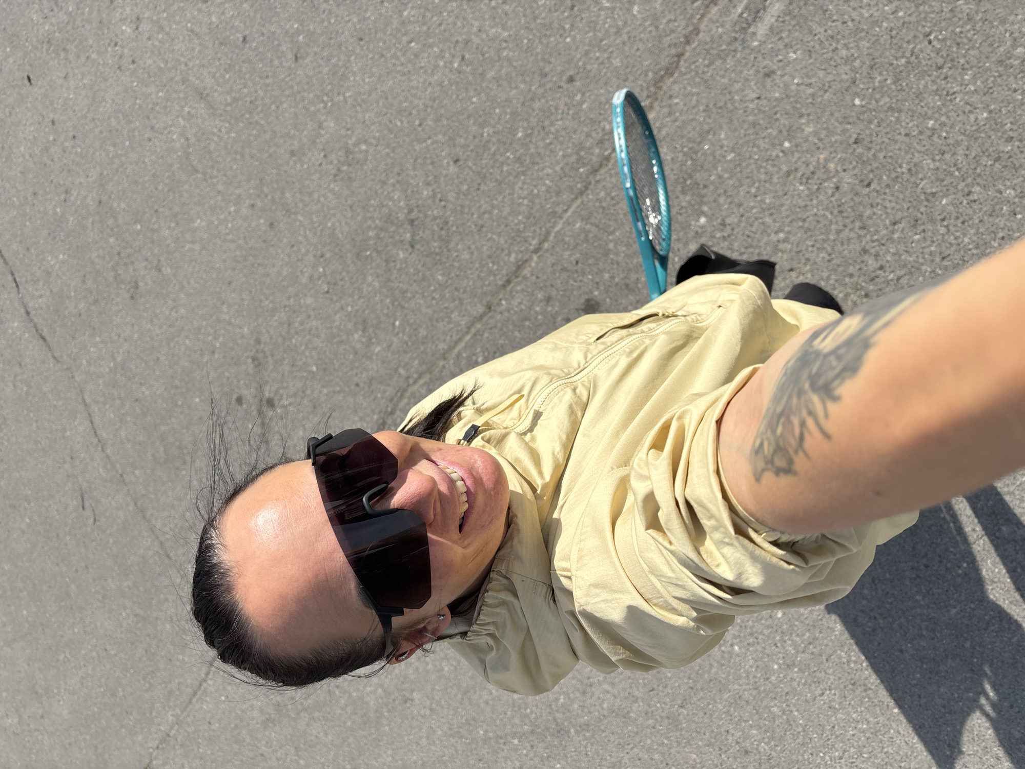 A person is taking a selfie outdoors. They are wearing dark sunglasses and a beige jacket. The person has a tattoo on their arm. In the background, there is a blue racquet and paved ground. No evidence in the image confirms the location as the city of Chaikovsky, Russia.