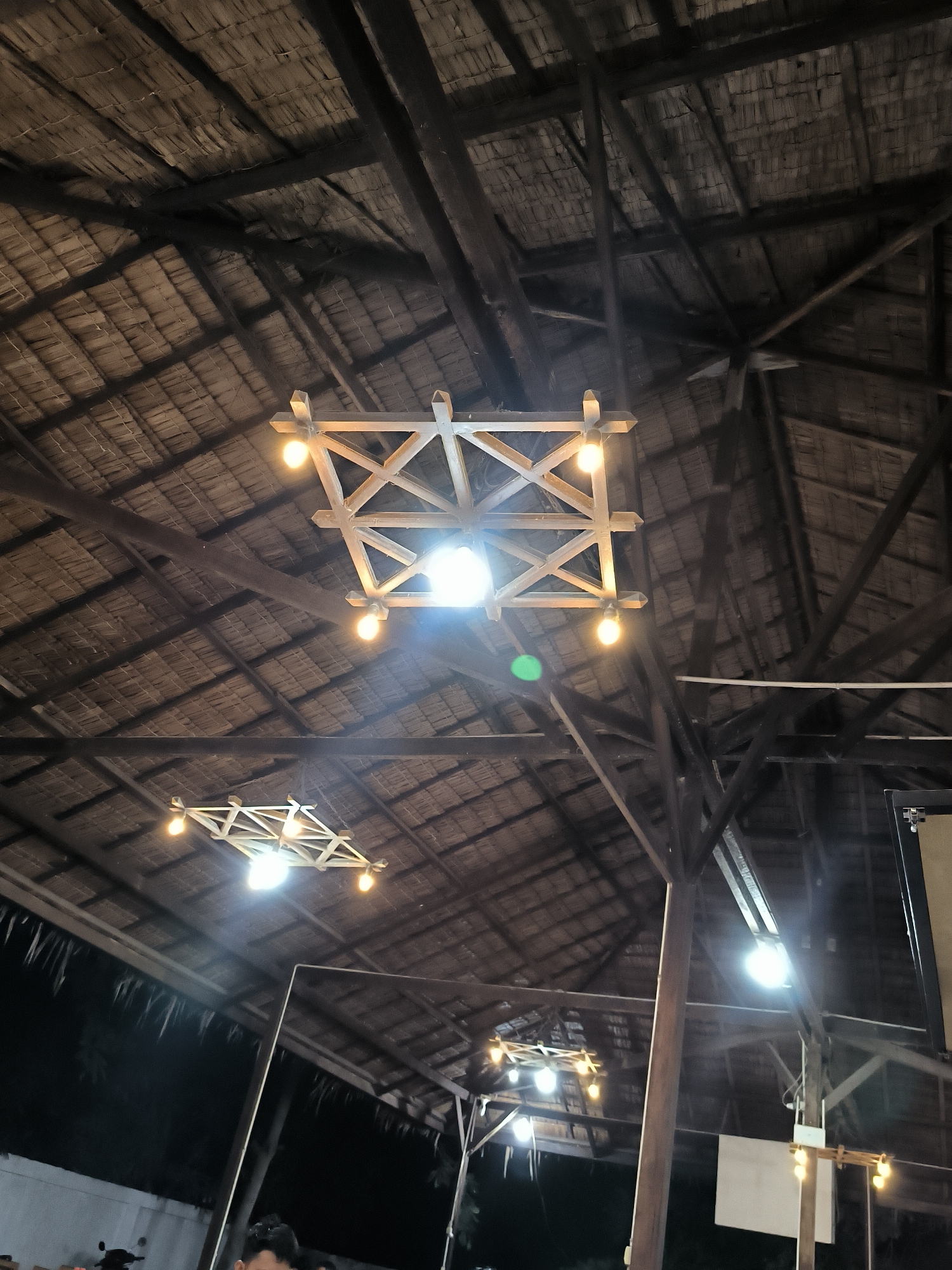 The image depicts the interior of a building with a thatched roof structure supported by dark wooden beams, presumably in Cempeudak, Indonesia. Three square wooden light fixtures with multiple exposed bulbs illuminate the space. Reflections suggest there are mirrored surfaces present. In the foreground, a portion of the building’s exterior is visible, with partial glimpses of people and vehicles. Additional lights are visible in the reflection, with one positioned closer to the wall.