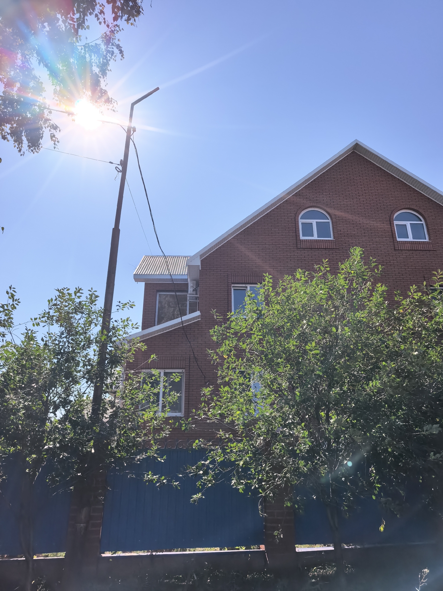 A two-story brick house with a metal roof stands under a clear blue sky. Two arched windows are visible on the upper floor of the house, and one rectangular window on the lower floor. A tree with green leaves partially obscures the view of the house. In the foreground, a blue fence runs along the property line. The sun is bright and creates lens flare in the image, suggesting it is daytime. There are no people visible in the image. The location is identified as Sterlitamak, Russia.