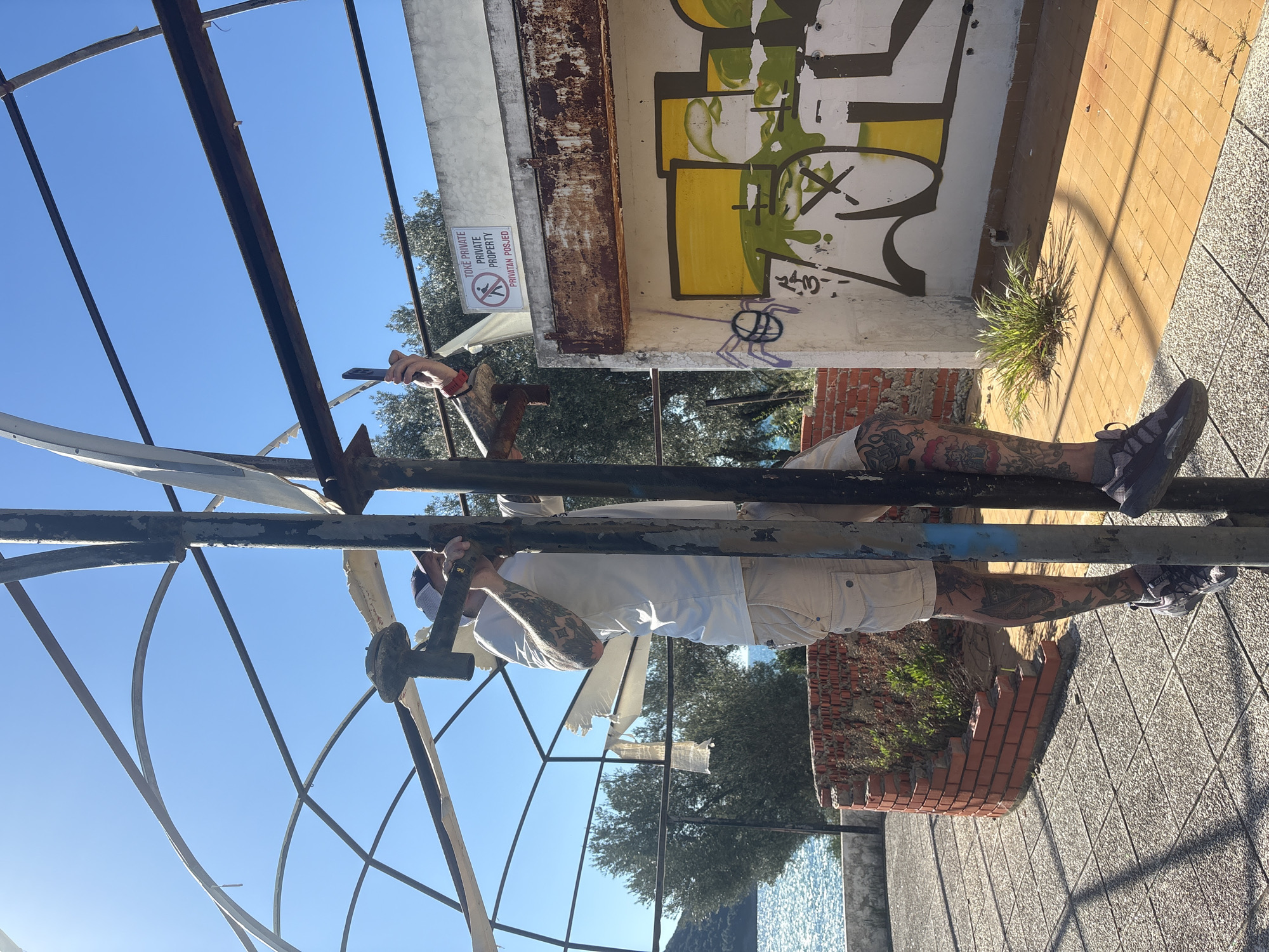 The image shows a man clinging to the rusted, exposed metal framework of a ruined structure in Bratica - Braticë, Montenegro. The man is wearing light-colored shorts and a white shirt. He has tattoos on his arms and legs. The structure is partly collapsed, with tattered white fabric hanging from some sections. Graffiti adorns a nearby concrete wall, and a "No Trespassing" sign is visible. Beyond the structure, trees are visible, and a glimpse of water appears at the bottom of the frame. The ground is paved with textured stone tiles, and a dilapidated brick wall borders a section of the ground. The sky is clear and blue.