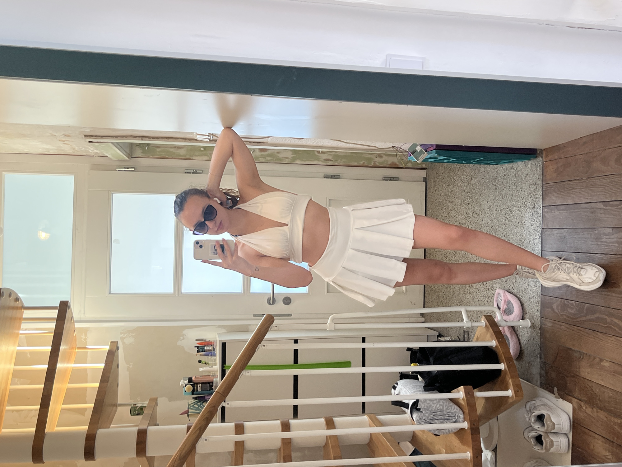 A young woman stands indoors, facing a mirror and holding a smartphone to take a selfie. She is wearing a white halter top and a matching pleated mini skirt. She is also wearing sunglasses and one earbud. She has one arm raised with her hand near her head. The background includes a staircase, a white door with glass panels, exposed wall construction, and wood flooring.
The location in the city of Saarbrücken, Germany cannot be determined from this image.